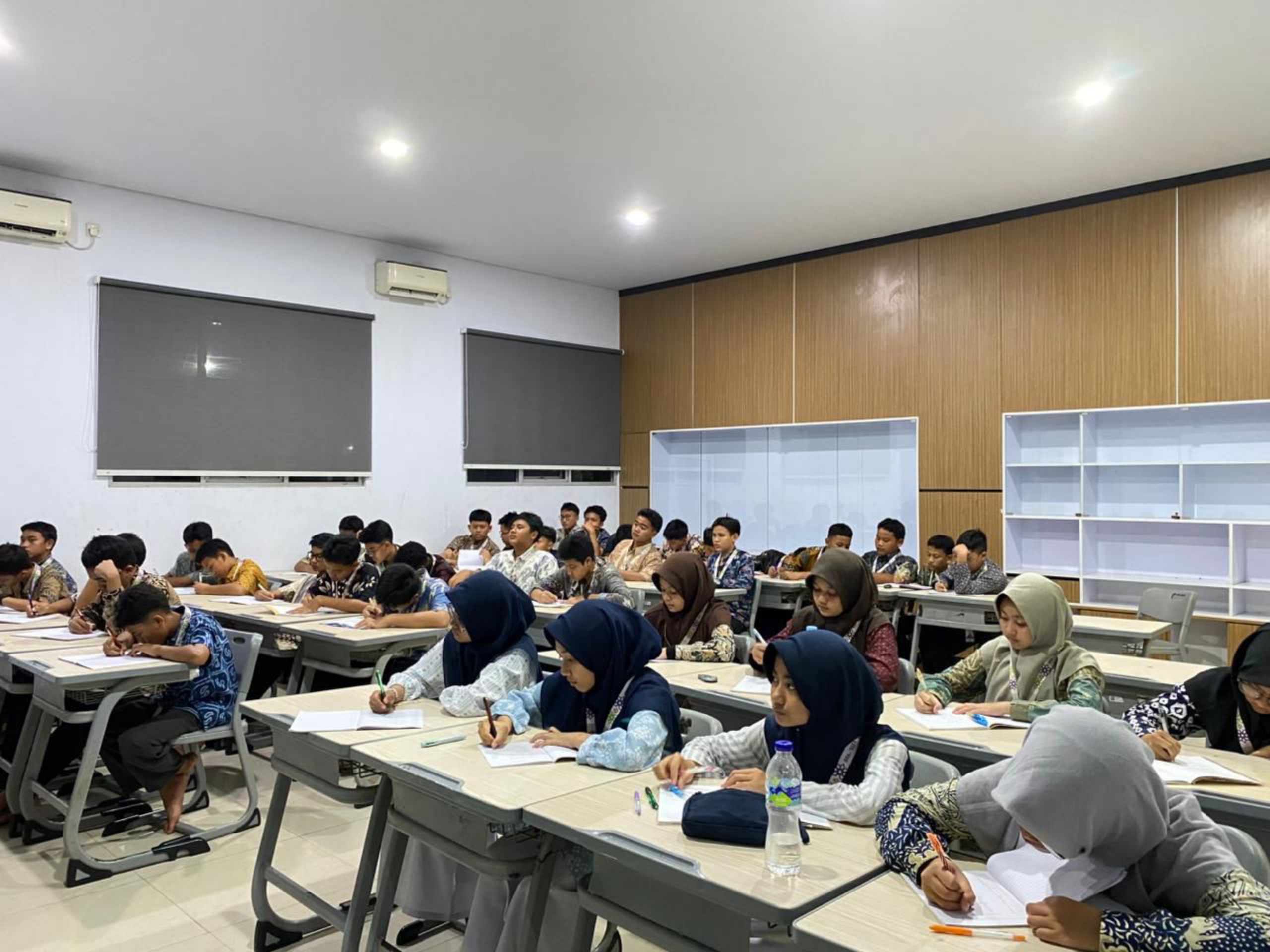 A classroom filled with many students seated in rows, concentrating on writing at desks while some girls wear hijabs, with bright ceiling lights, grey window blinds and a wood‑panelled wall behind them.