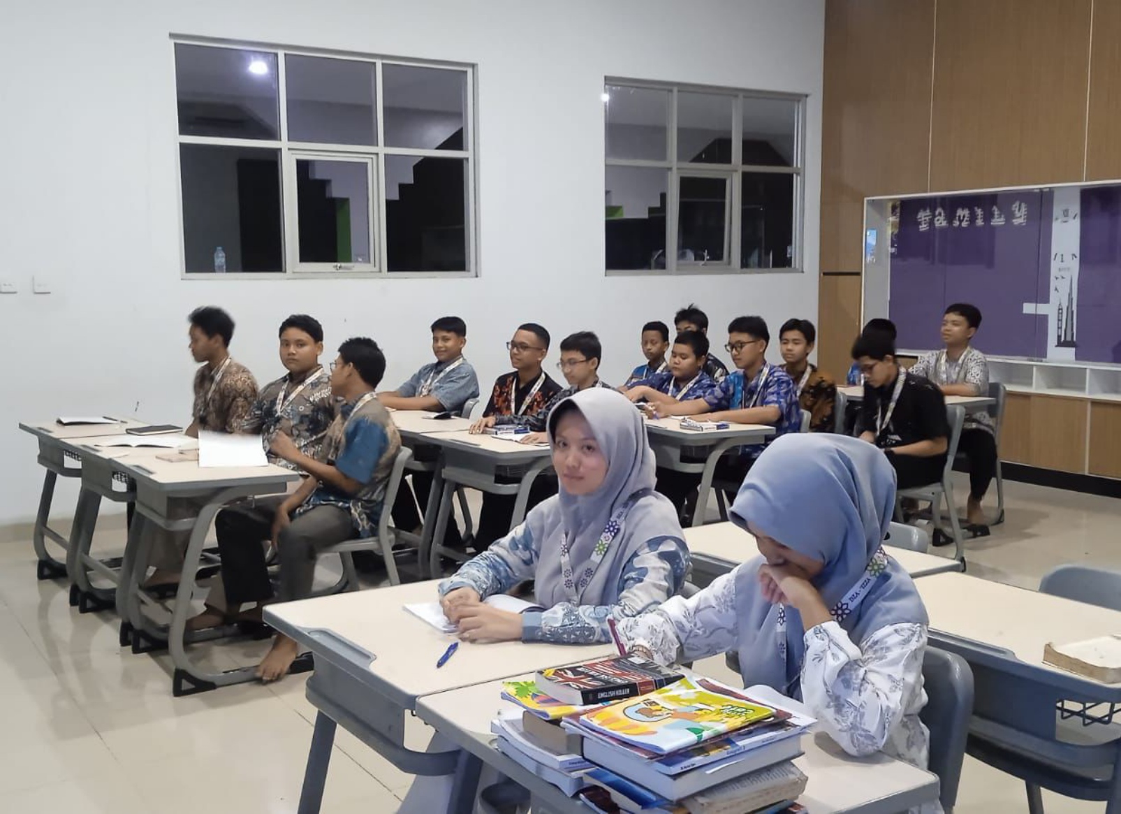 A classroom of teenage students seated at rows of desks, with two girls in light grey hijabs and school uniforms at the front desk surrounded by textbooks and pens and a group of boys in patterned shirts sitting behind them against a white wall with windows.