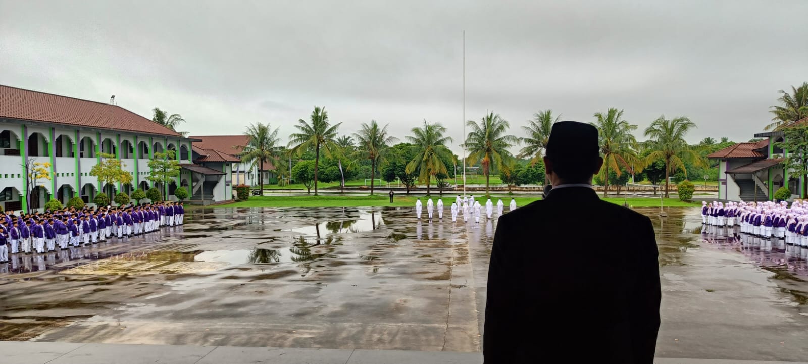 Silhouetted figure in a dark suit and cap stands on a covered platform overlooking rows of students in purple-and-white uniforms lined up on a wet school courtyard with palm trees and two-storey buildings under an overcast sky.