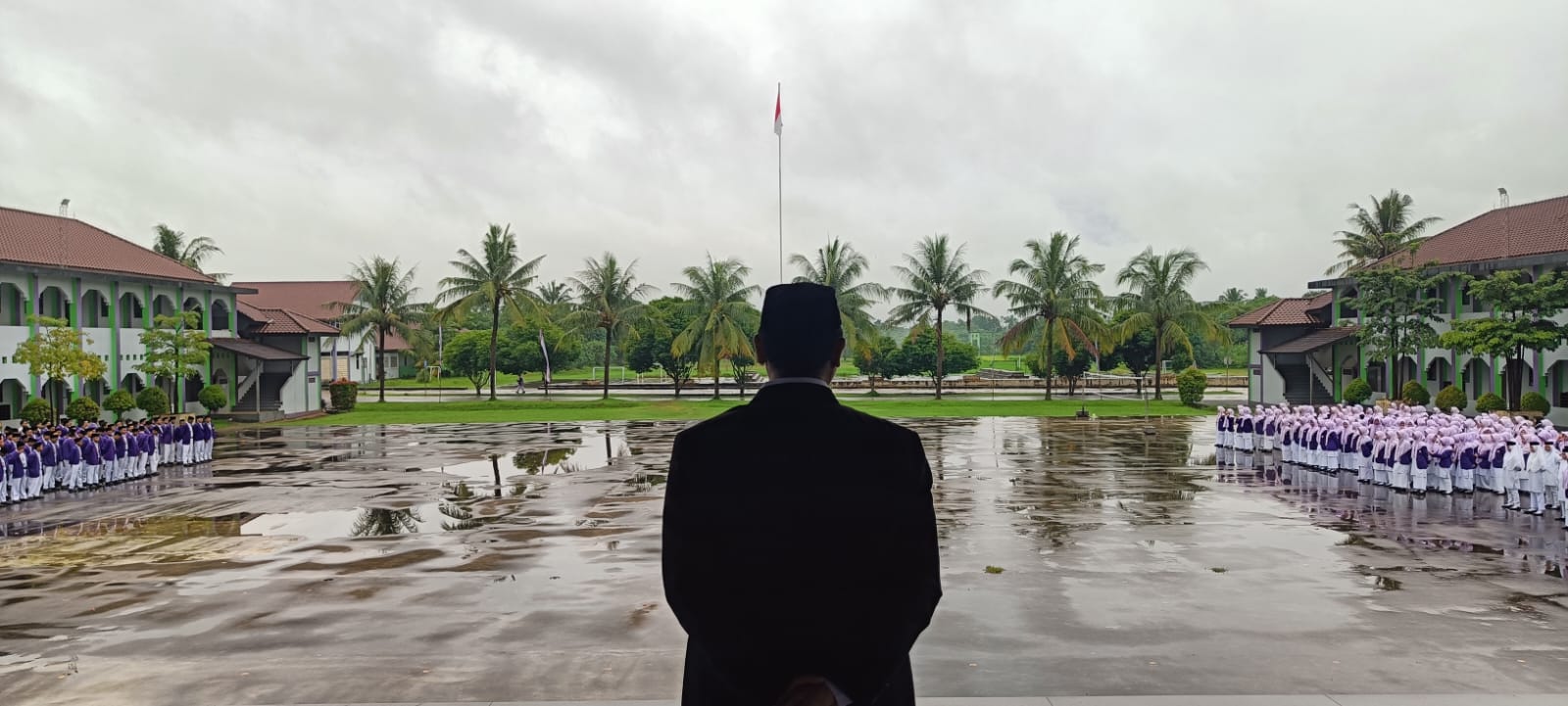 A man in a dark suit and black cap stands with his back to the camera overlooking a wet, reflective school courtyard where rows of students in purple and white uniforms line both sides facing a tall flagpole, with palm trees and two-storey school buildings framing the scene under an overcast sky.