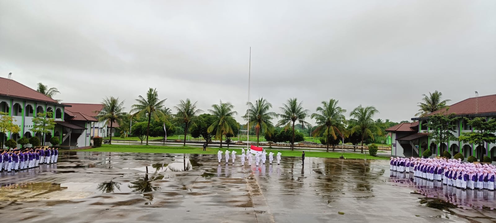 Students in purple-and-white uniforms stand in lines on a rain-wet school courtyard while a small colour guard in white raises a red-and-white flag at the central flagpole, palm trees and green-trimmed school buildings reflected in puddles under an overcast sky