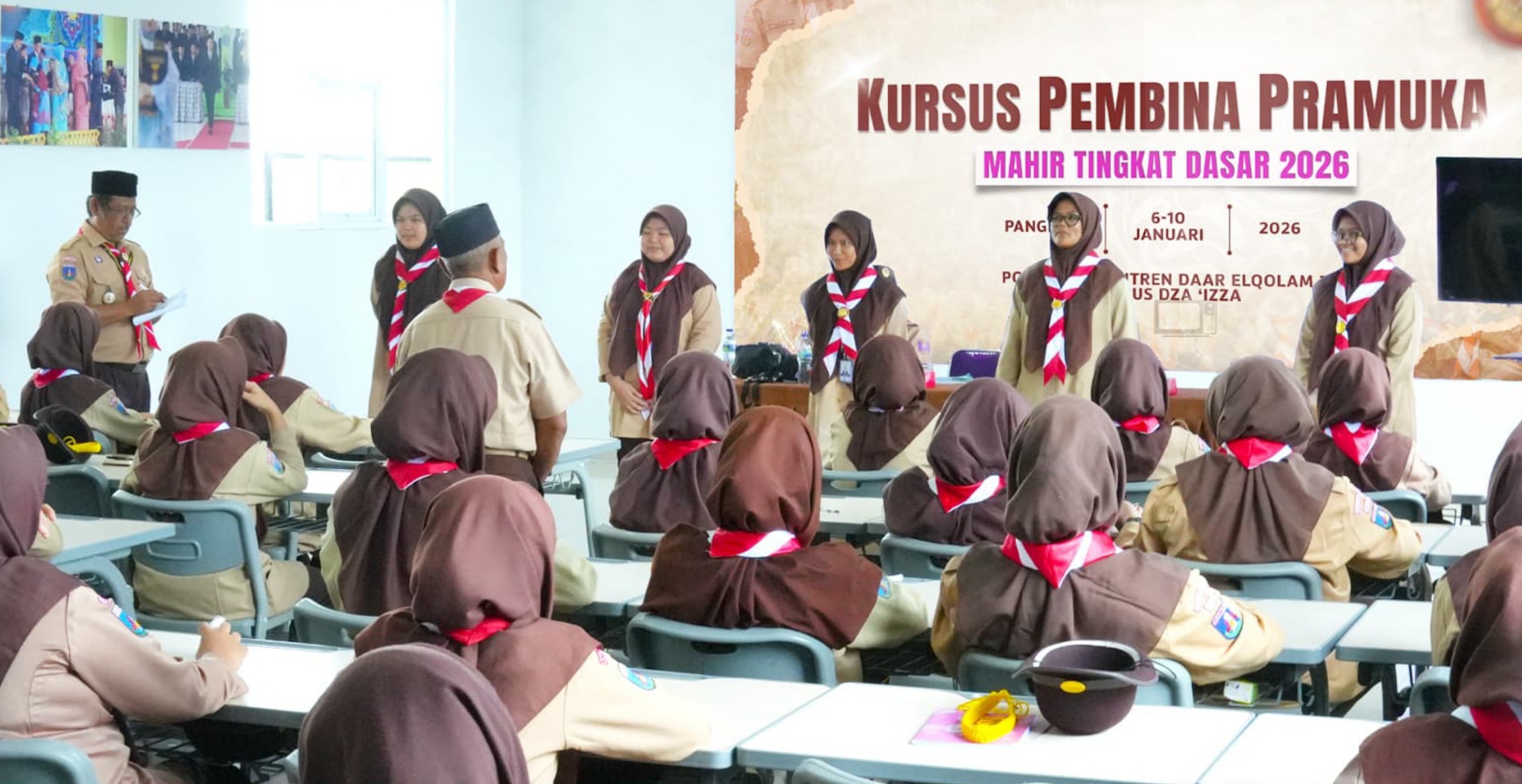 Rows of female scout trainees in brown headscarves and tan uniforms with red-and-white neckerchiefs sit at desks facing a panel of instructors and a large banner reading 'Kursus Pembina Pramuka Mahir Tingkat Dasar 2026' in a classroom.