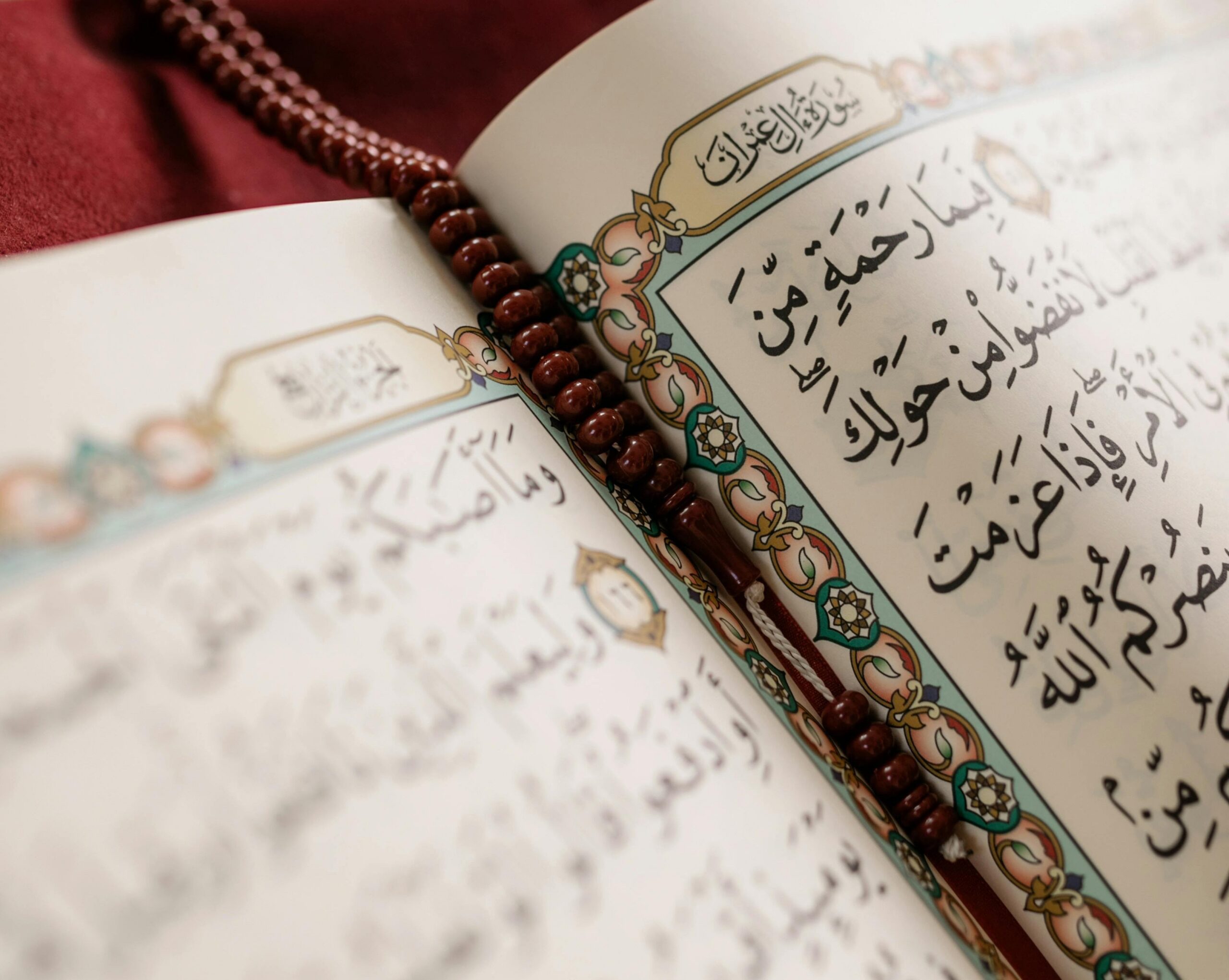 Open Qur'an showing Arabic calligraphy and an ornate turquoise-and-gold border, with a strand of polished brown prayer beads resting along the pages on a soft red background.