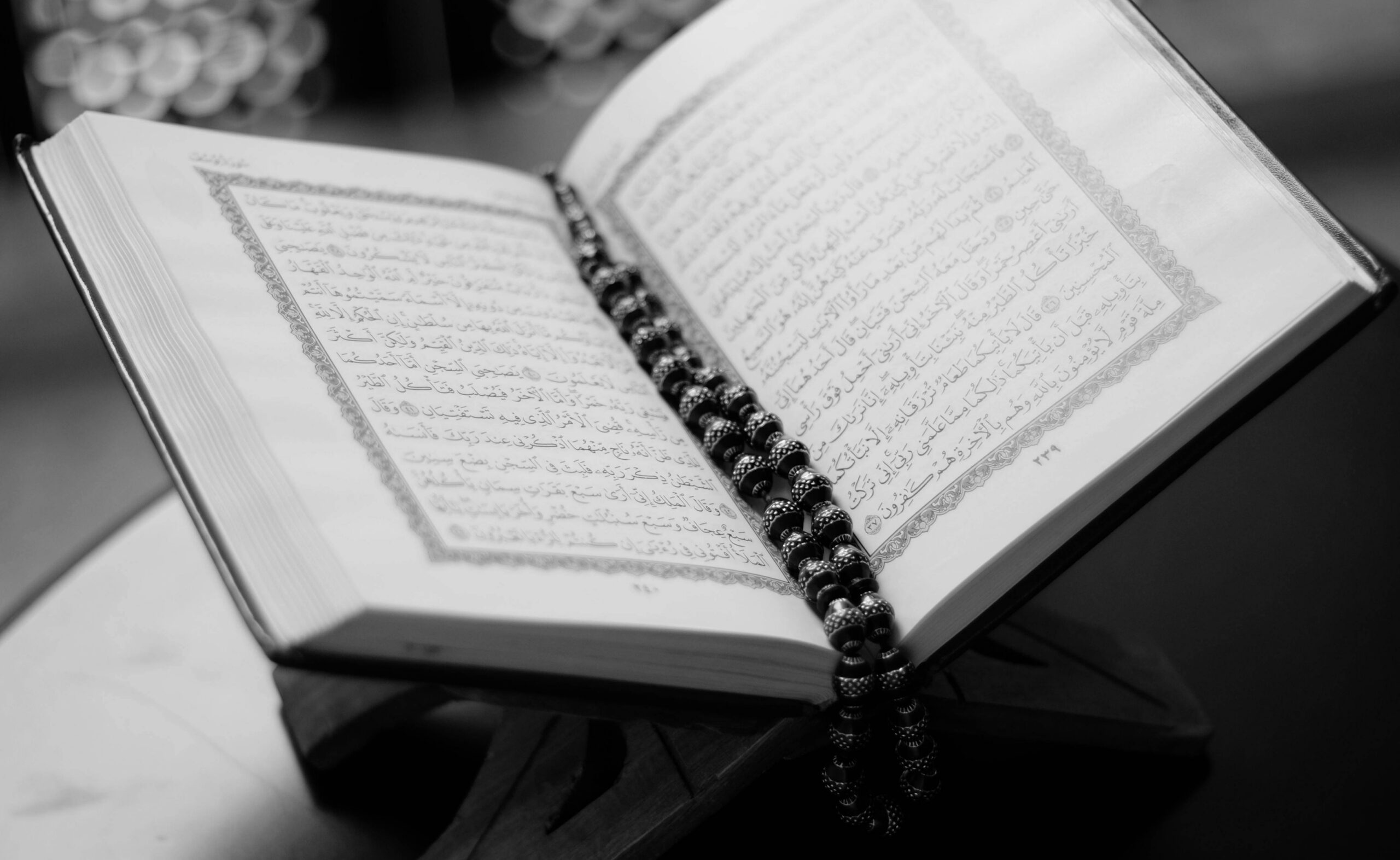 Black-and-white photo of an open Quran showing Arabic script and decorative page borders on a wooden stand, with a string of prayer beads draped along the centrefold.