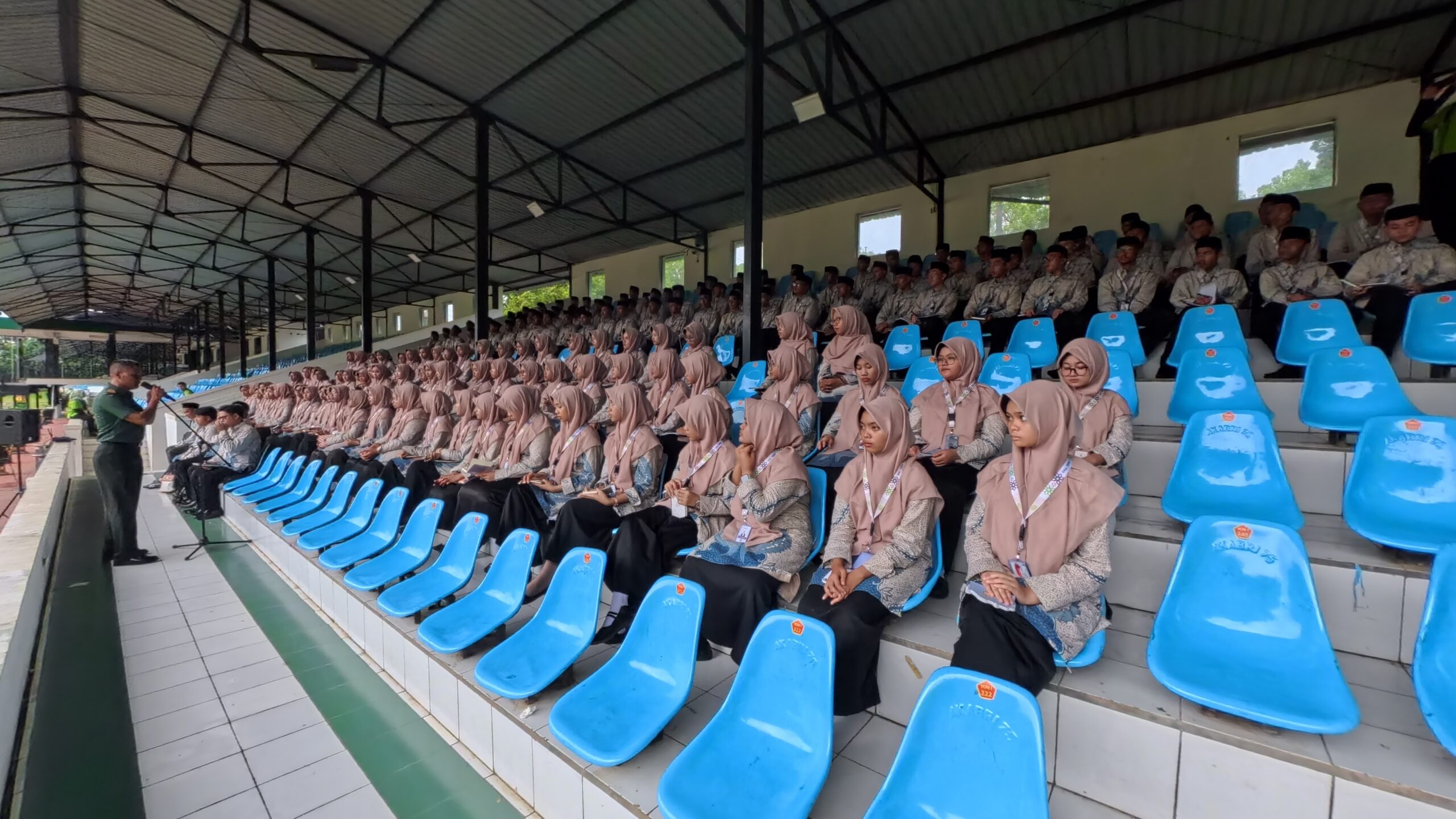 Rows of young women in beige hijabs and patterned uniforms seated on blue stadium chairs as a man speaks into a microphone.