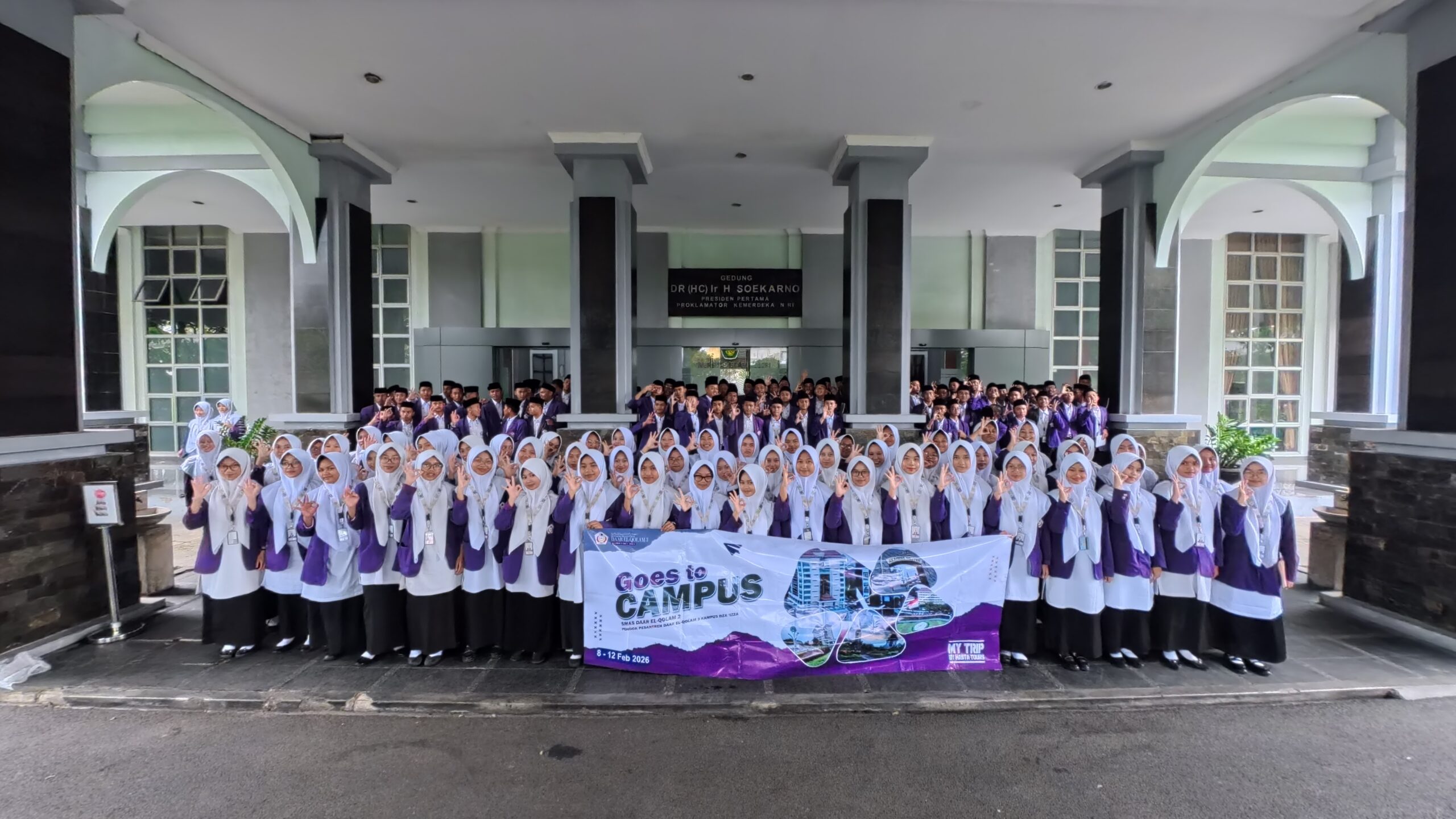 Large group of students, many in white hijabs and purple blazers, holding a Goes to Campus banner under a campus entrance.