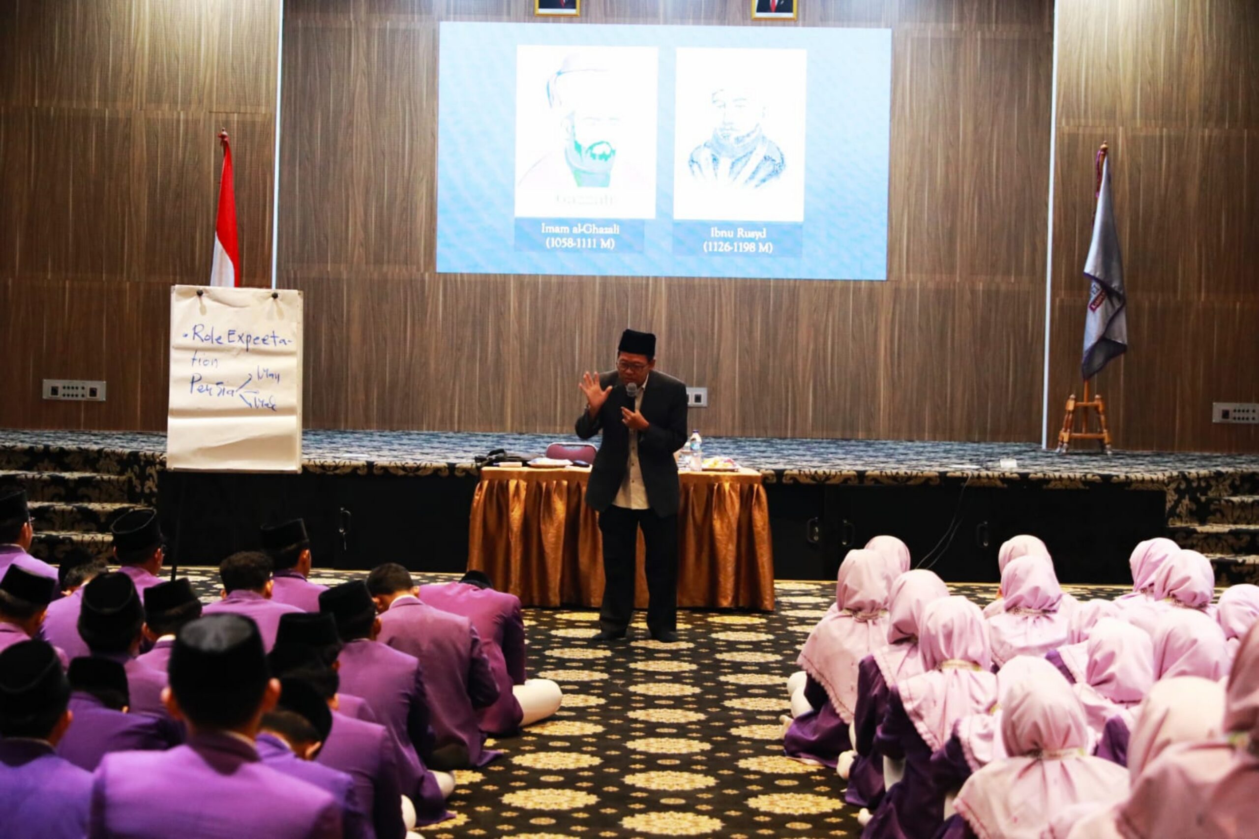 A male speaker in a black cap addresses rows of students in purple uniforms, with girls in pink hijabs seated on the right.