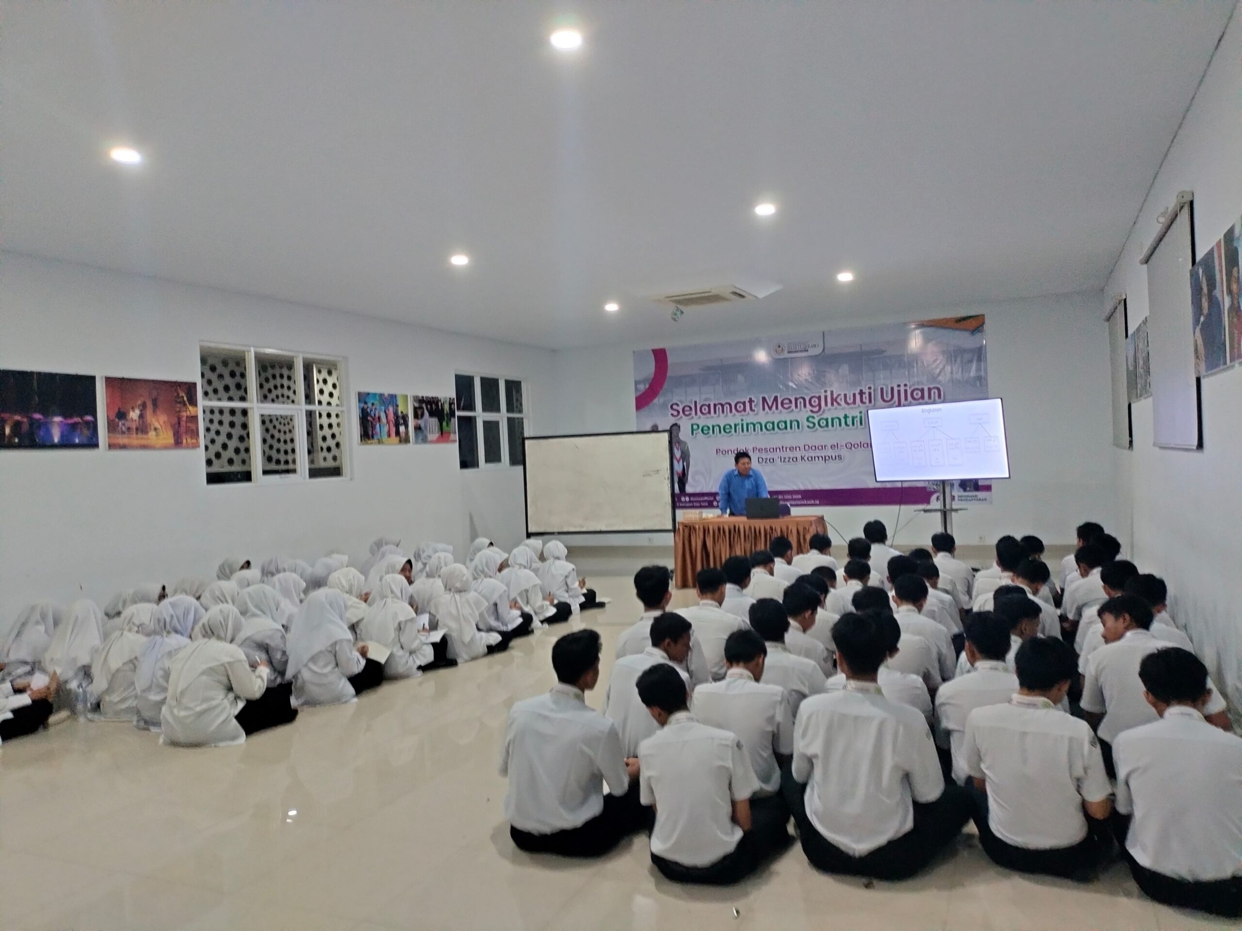 Students seated on the floor—girls in white hijabs at left, boys in white shirts at right—listening to a teacher at the front beside a banner and screen
