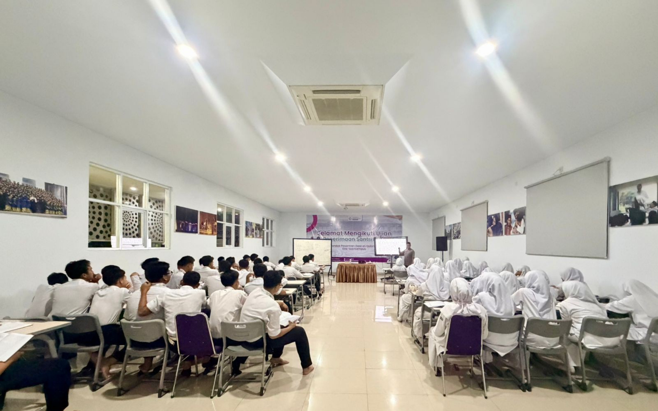 Bright classroom with rows of uniformed students—boys on the left, girls in white headscarves on the right—listening to a teacher at the front