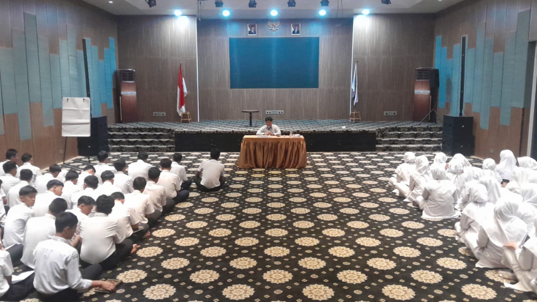 Students in white uniforms seated on patterned carpet in two groups facing a speaker at a table onstage with flags.