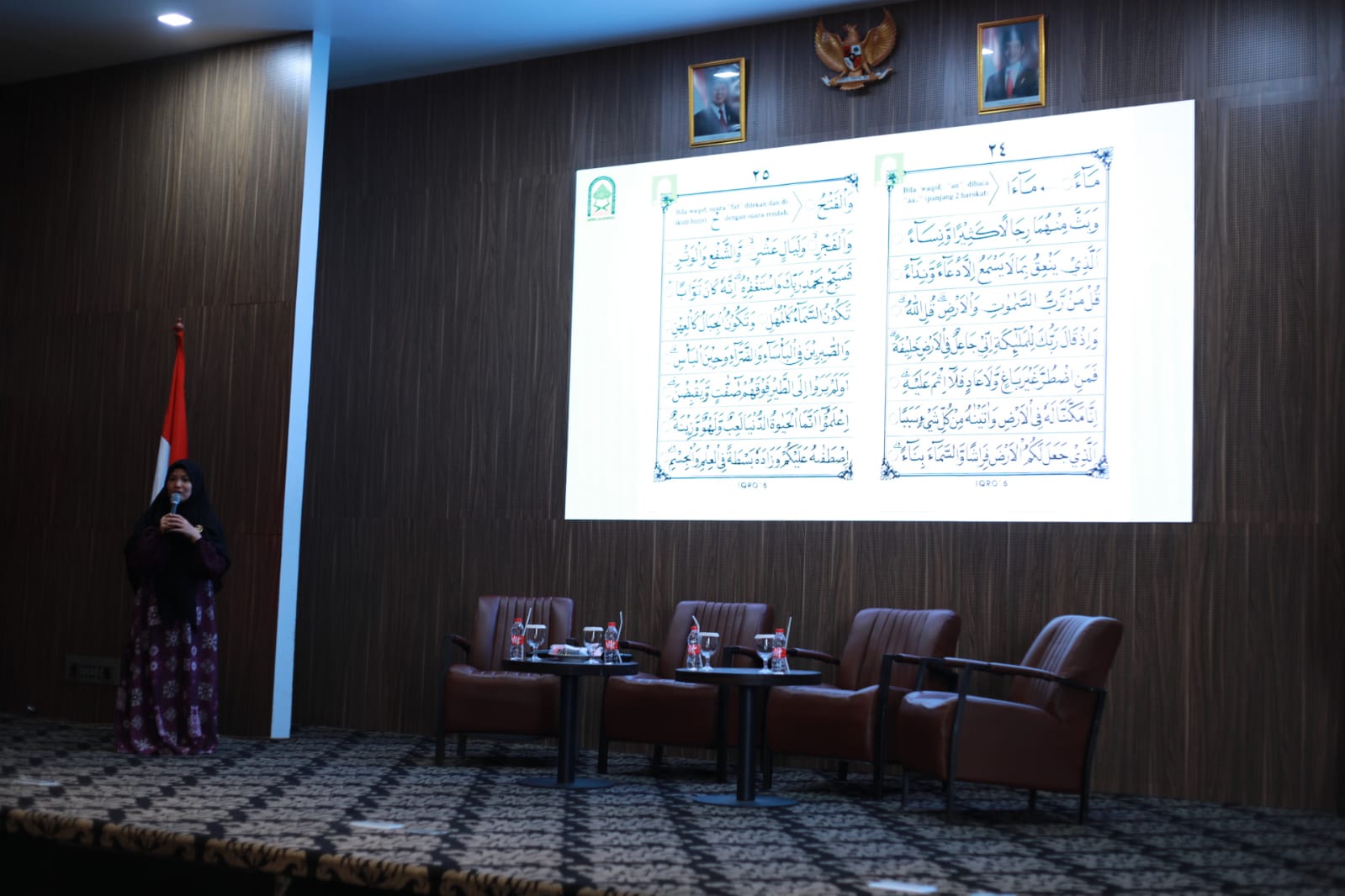 Woman in hijab speaking beside a large projected Arabic text on a stage, empty panel chairs, national flag and emblem above.