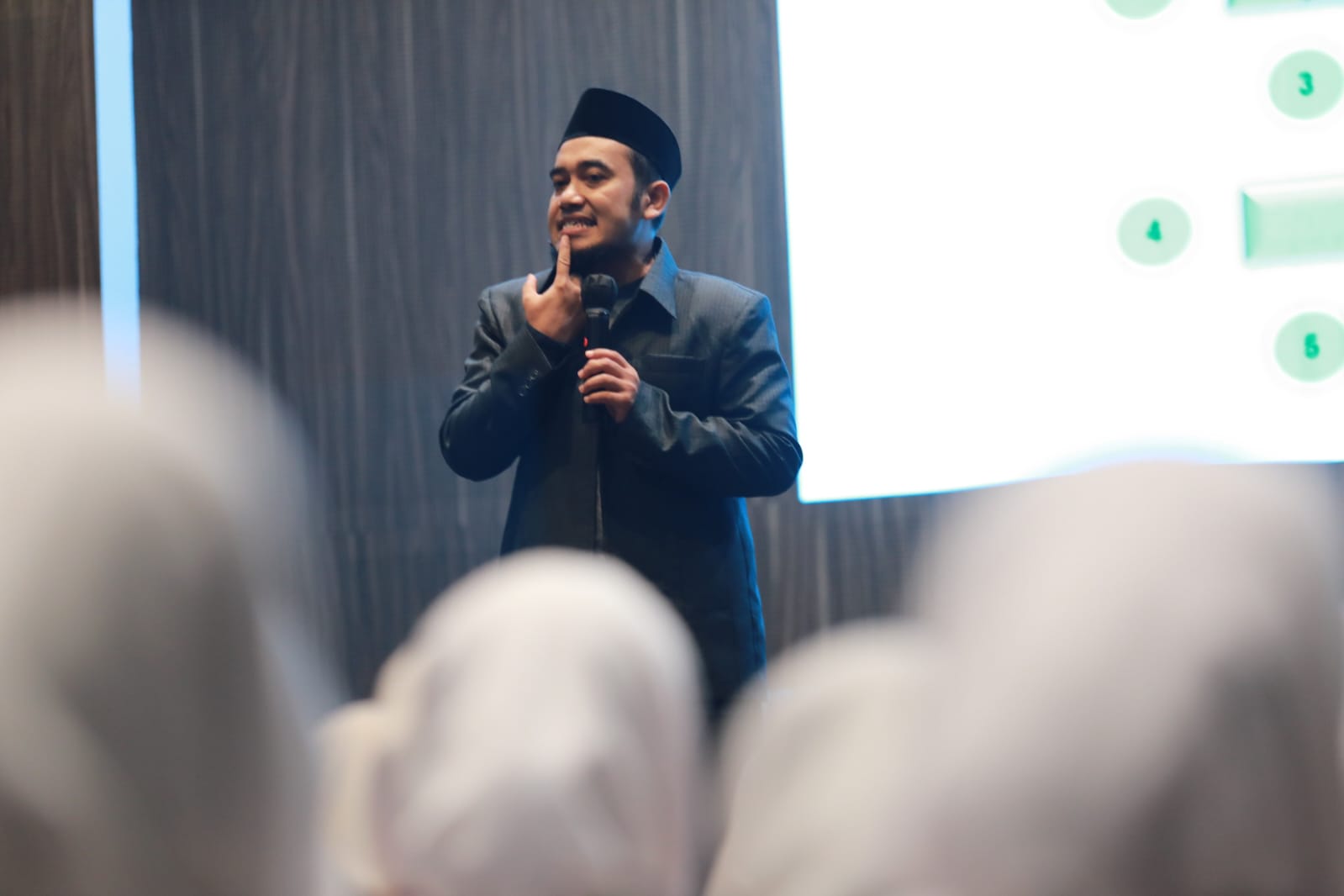 Man in dark jacket and black cap speaking into a microphone and gesturing to his face before a seated audience in headscarves