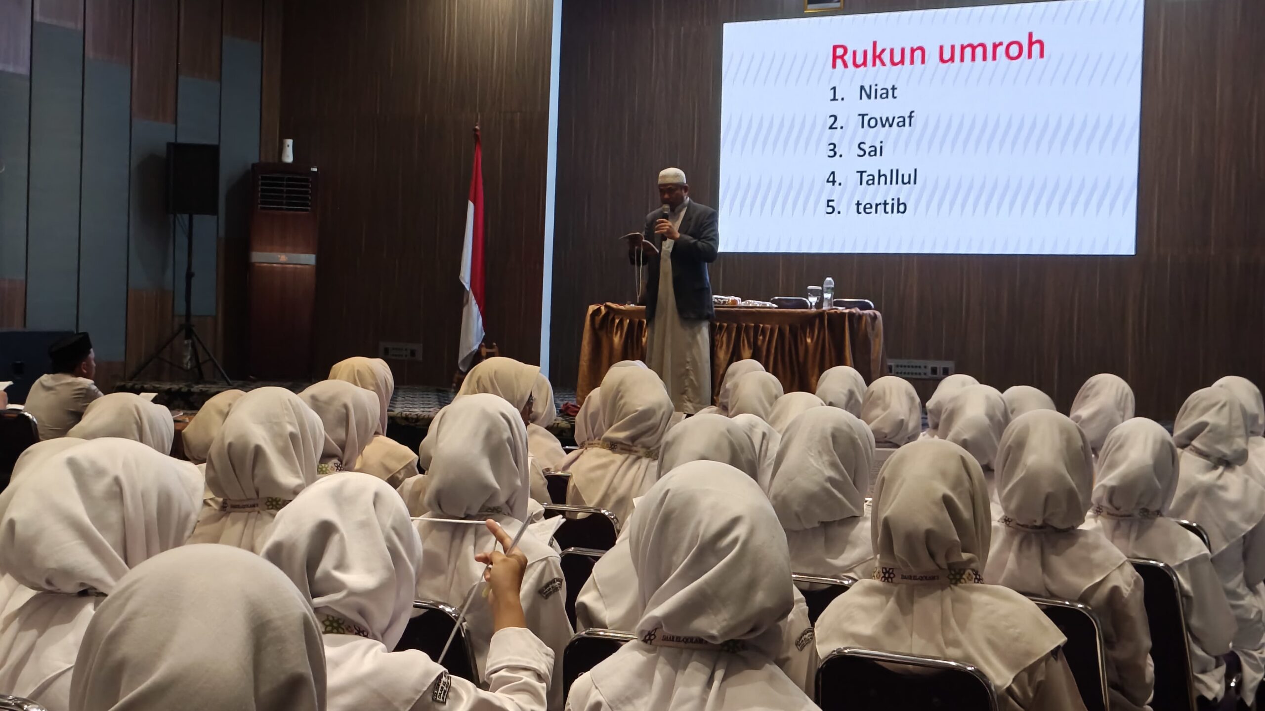 Women in white hijabs seated in rows listening to a male presenter on stage with a slide titled Rukun umroh