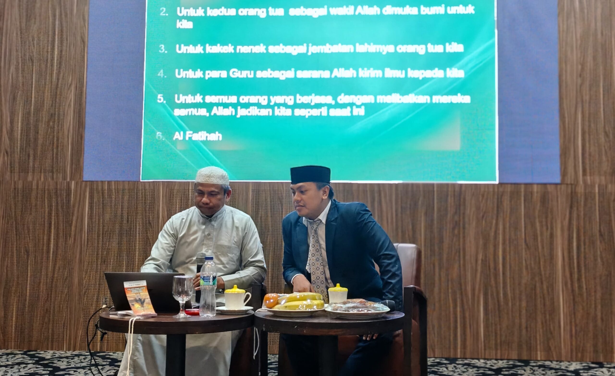 Two men on stage—one in white robe and cap, the other in suit wearing a black cap—with laptop and drinks before green screen.