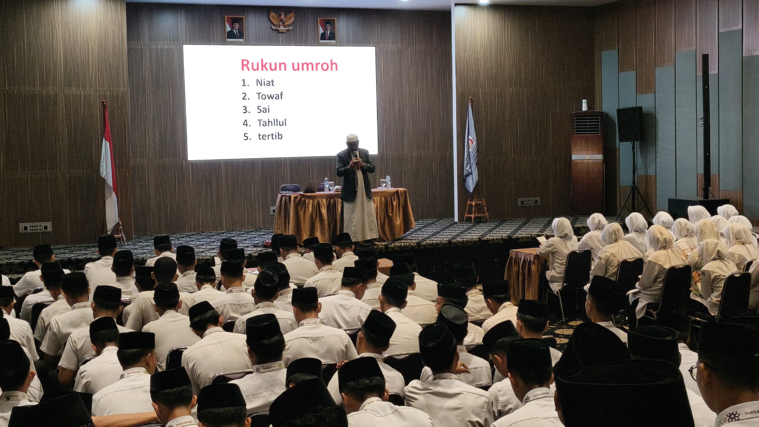 Speaker in robe addressing men in white shirts and black caps and women in white headscarves, slide reads 'Rukun umroh'.