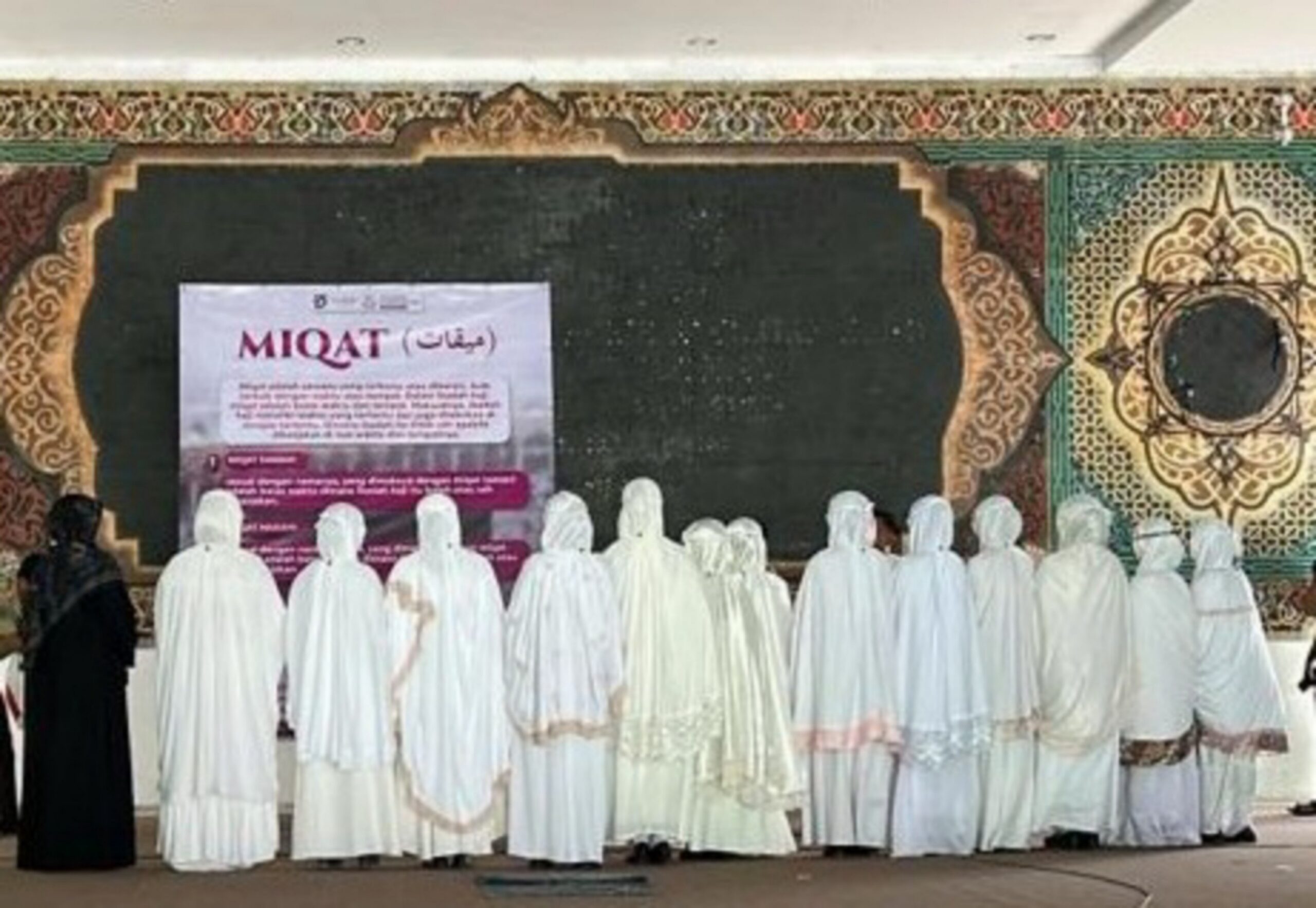 Women in white prayer robes standing in a row facing an ornate wall with a large black panel and a MIQAT poster.