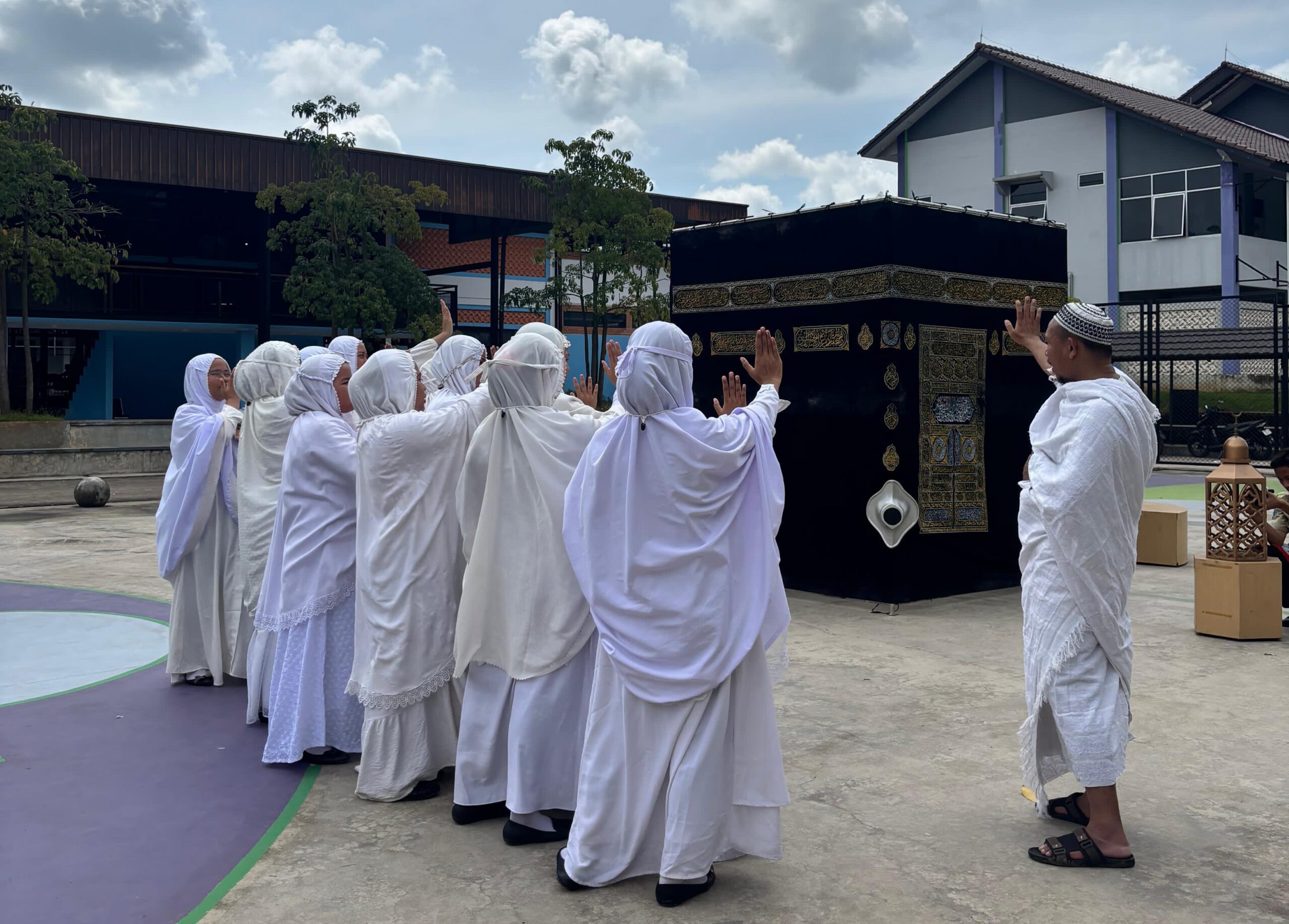 Women in white robes and headscarves practising tawaf gestures around a black Kaaba replica, guided by a man in white ihram.