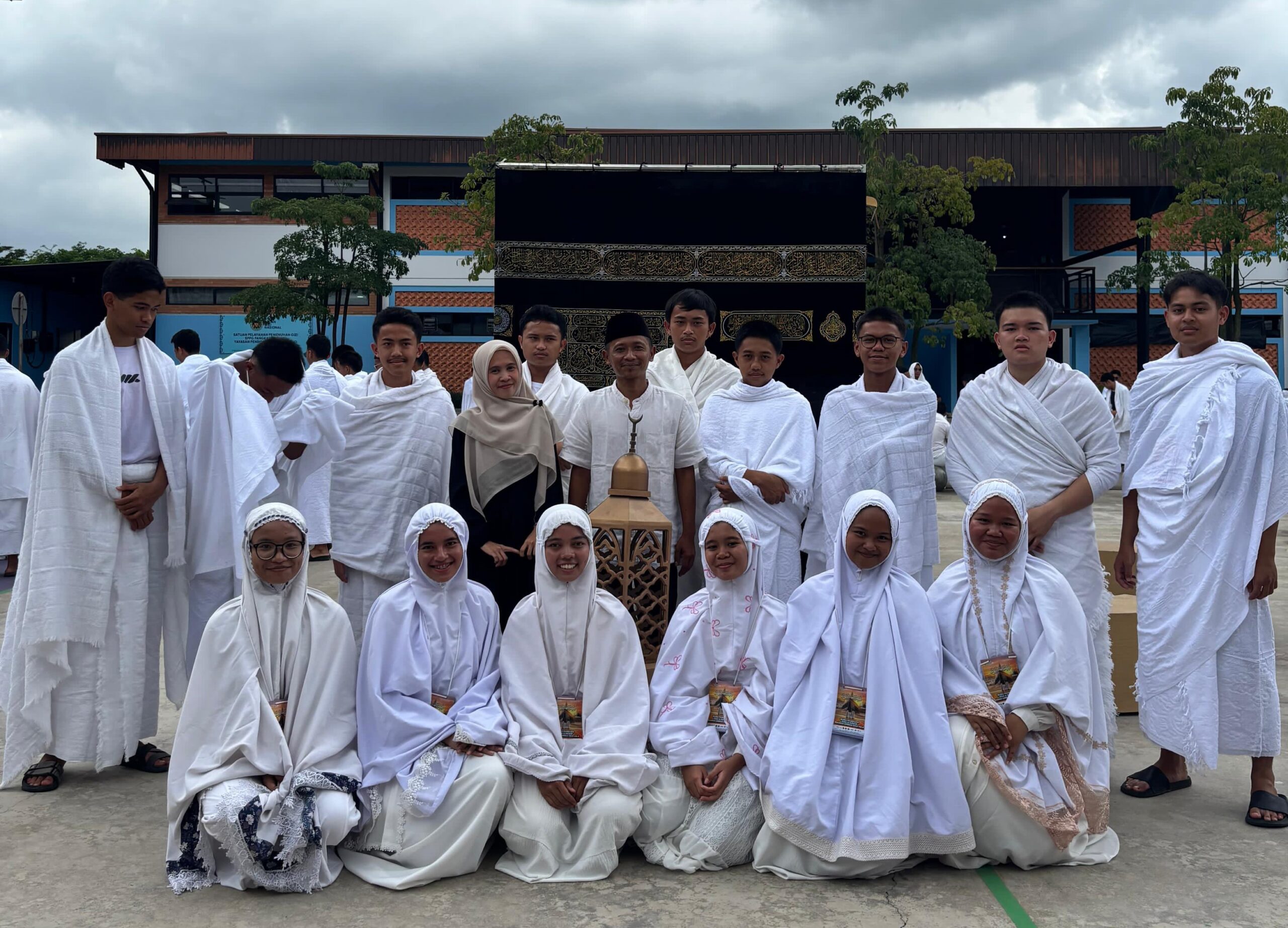 Group of students in white pilgrimage garments posing in front of a black Kaaba replica at school under cloudy sky.