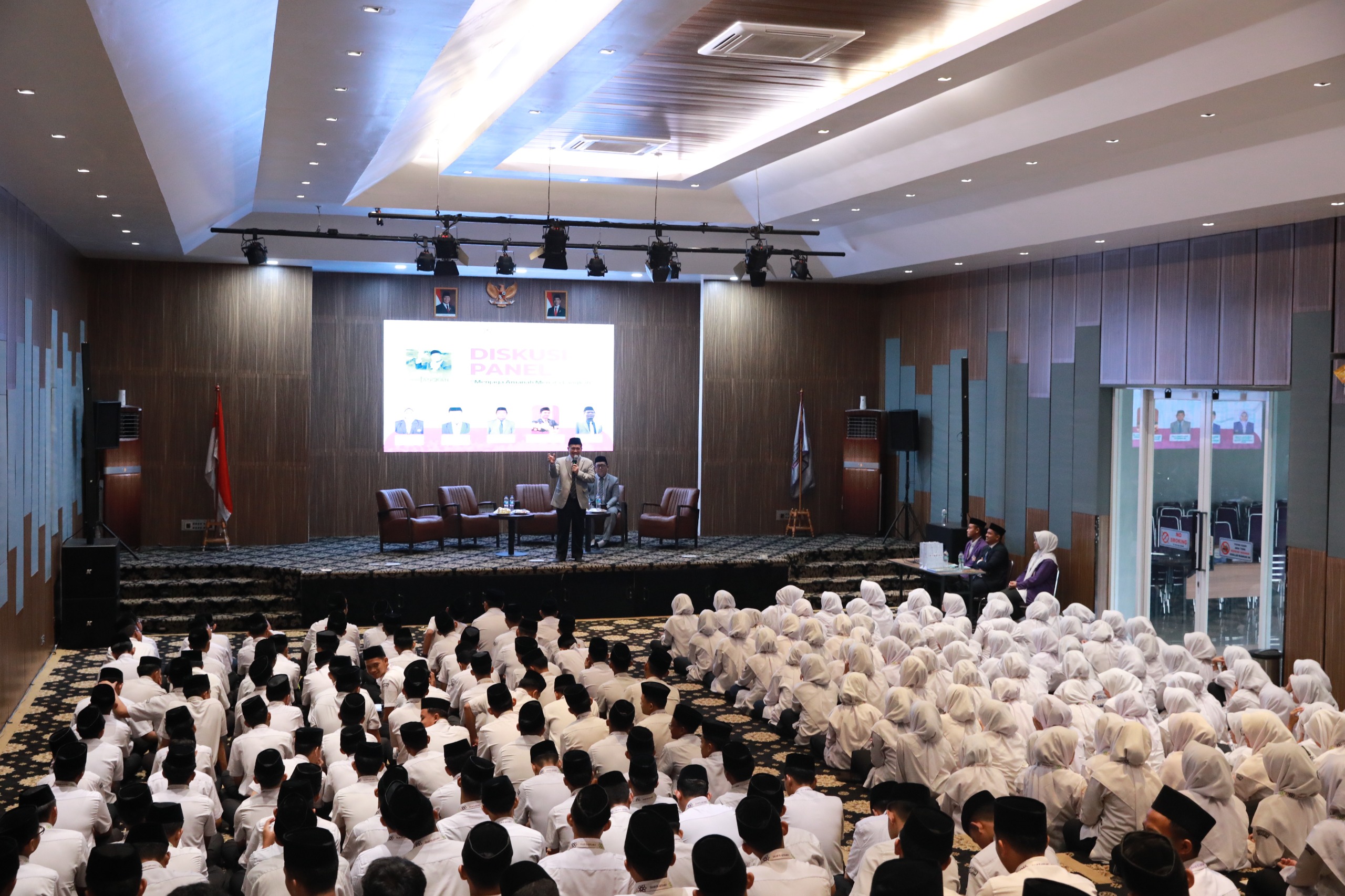 Students in white uniforms—boys with black caps and girls in hijabs—seated on the floor, listening to a speaker on a stage with a projection screen.
