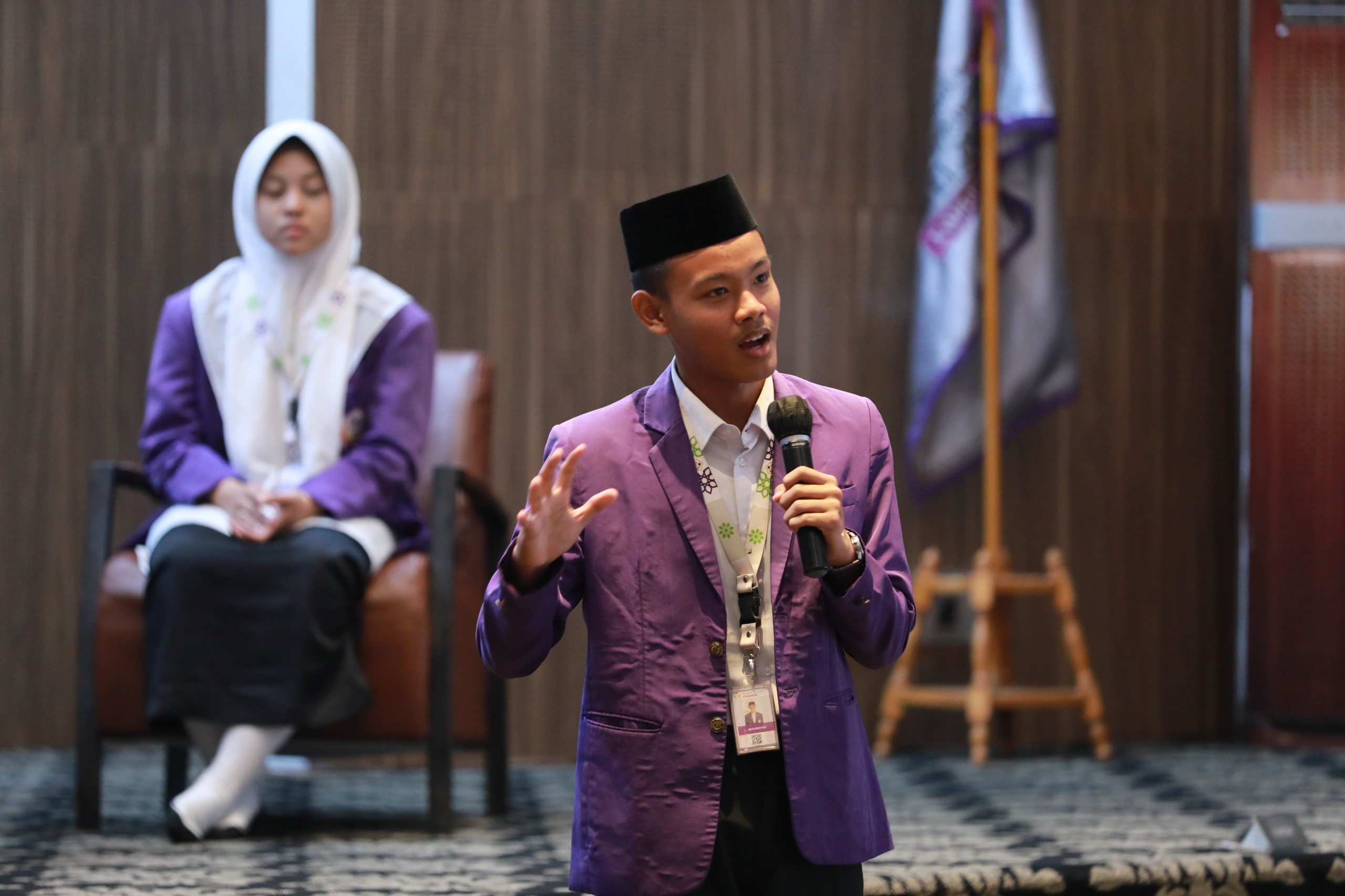 Young man in purple blazer and black cap speaking into a mic at a conference, a seated woman in a white hijab behind him.