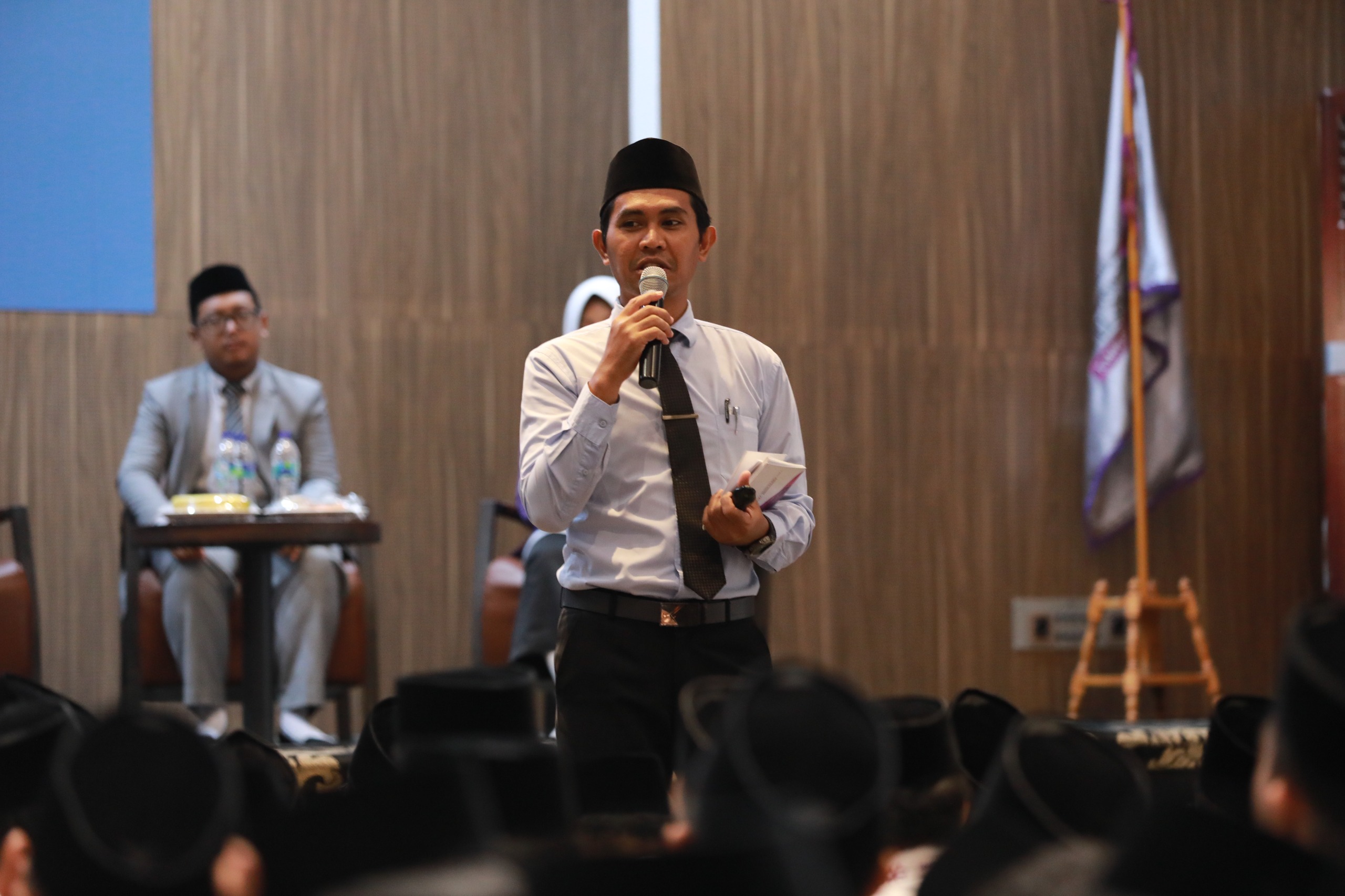 Man in white shirt, black tie and traditional cap holding a microphone and notes speaking to an audience in a formal hall.