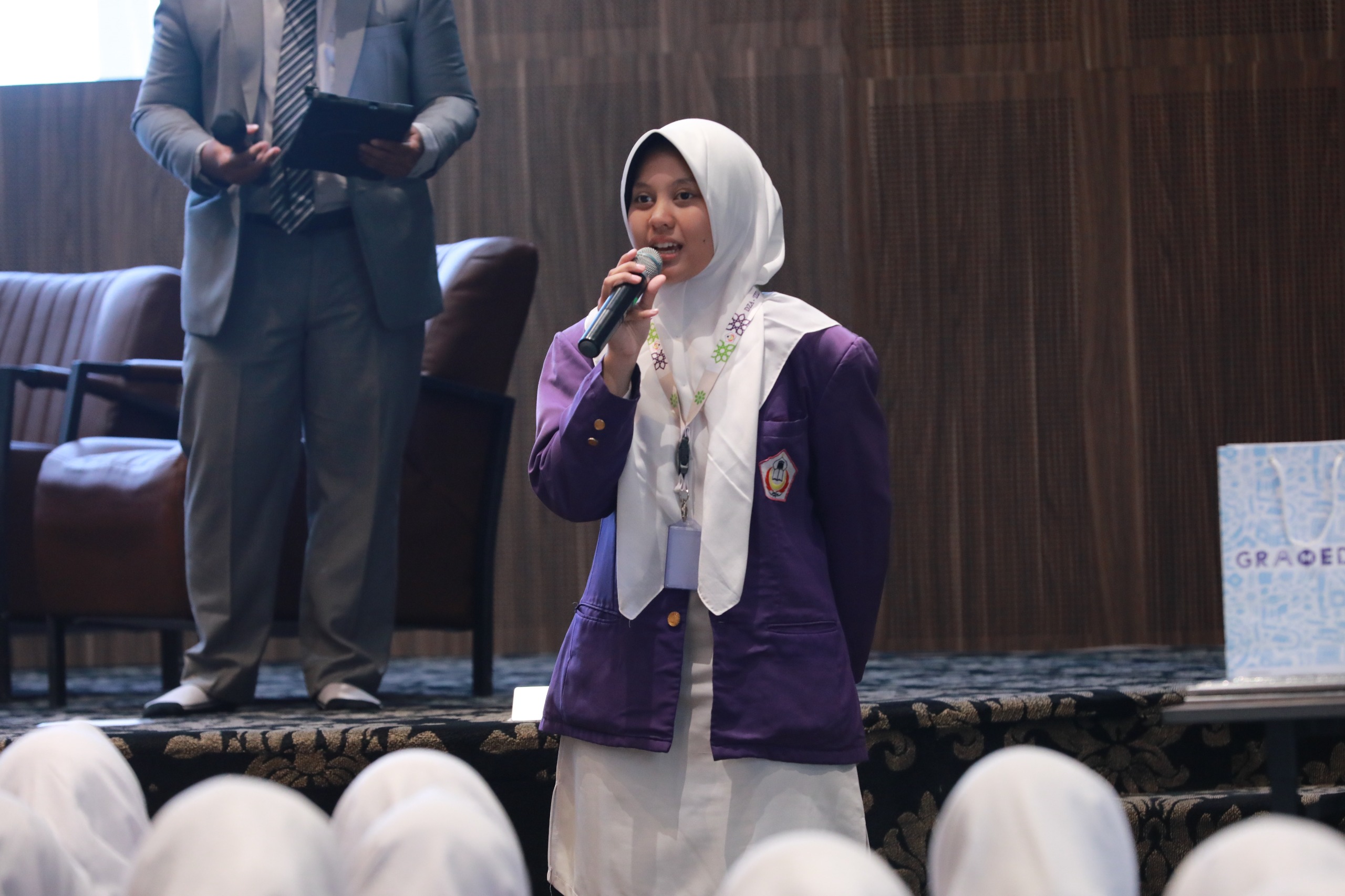 Teenage woman in white hijab and purple blazer speaking into a microphone on stage with a suited host standing behind her