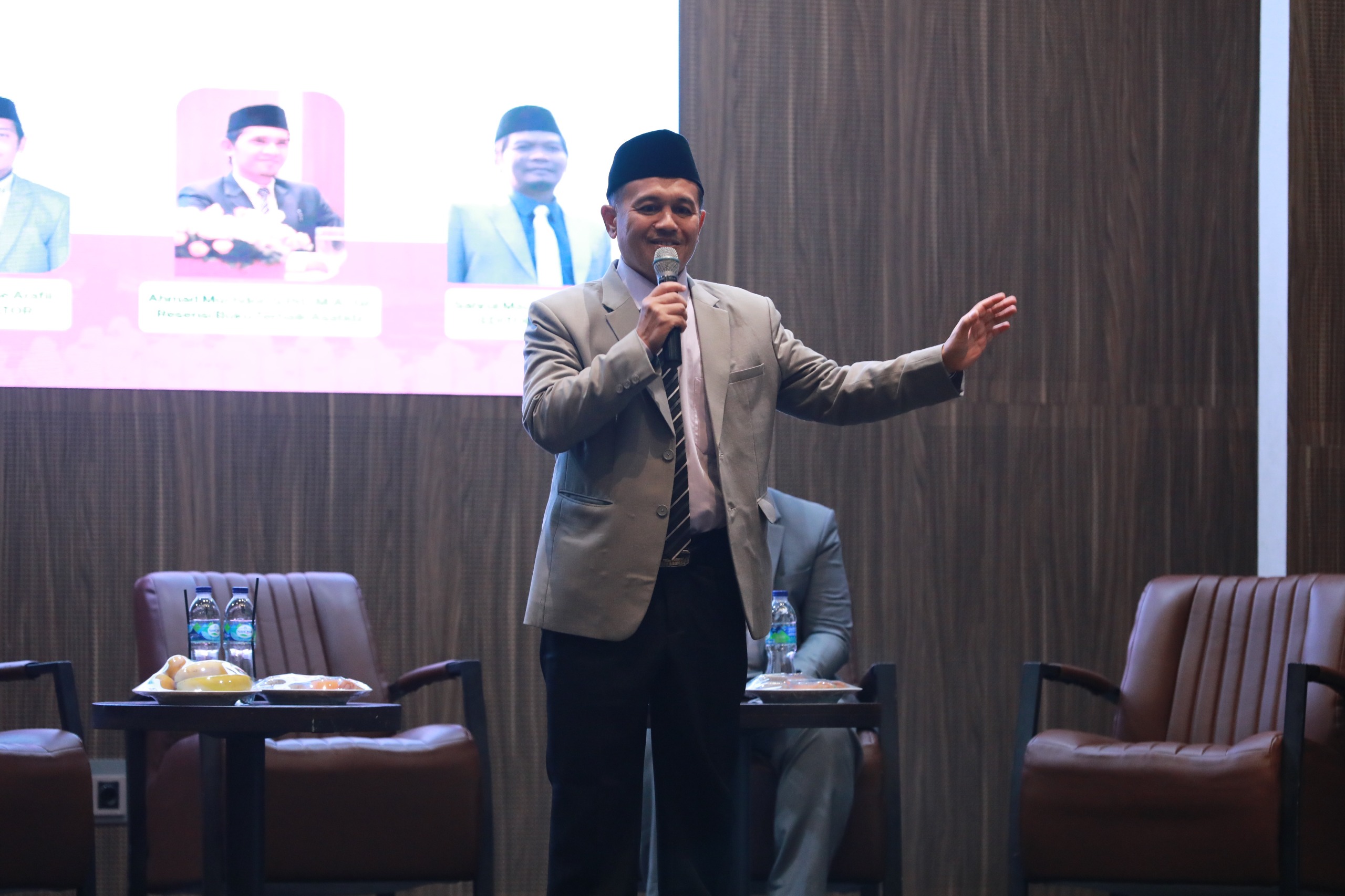 Man in grey suit and black cap holding a microphone, gesturing on stage beside chairs and a table with water and snacks.