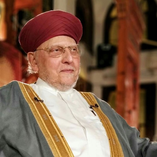Elderly man in a maroon turban and glasses wearing a white shirt and gold-trimmed grey robe, seated in an ornate interior.