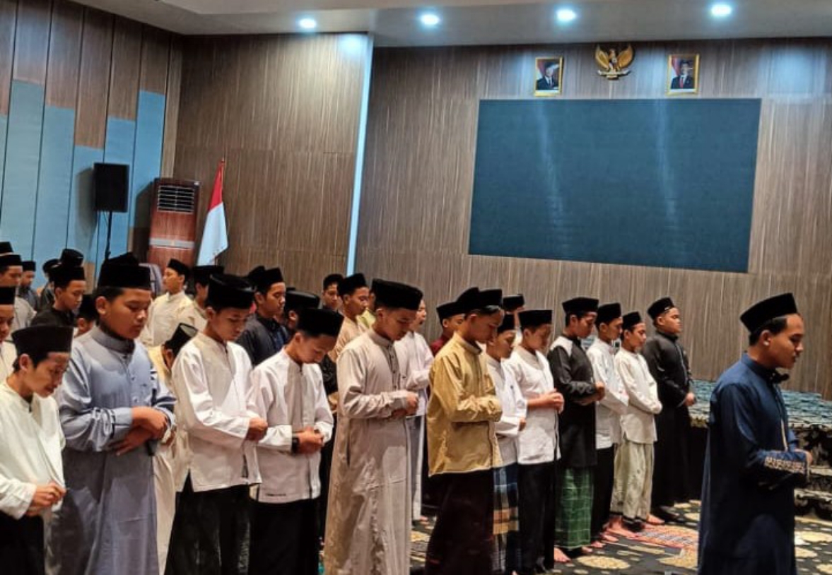 Rows of boys and young men in traditional robes and black caps praying in a hall with an Indonesian flag.
