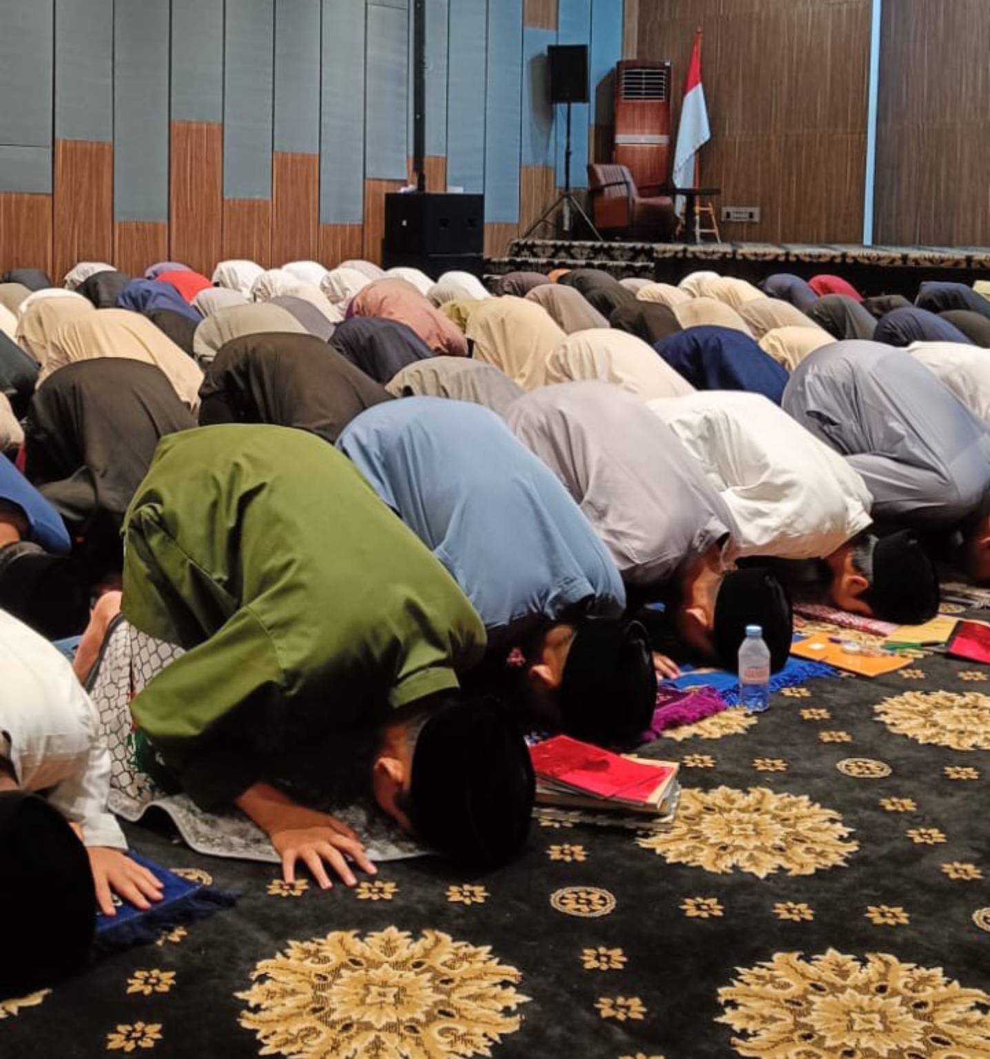 Men praying in prostration on a patterned carpet in a hall, with books and a water bottle beside them and a red-and-white flag on stage.