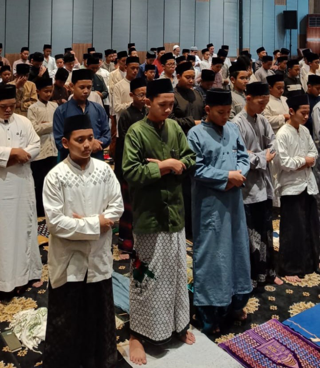 Rows of boys and young men in songkok caps, sarongs and long shirts standing with arms folded in a prayer hall.