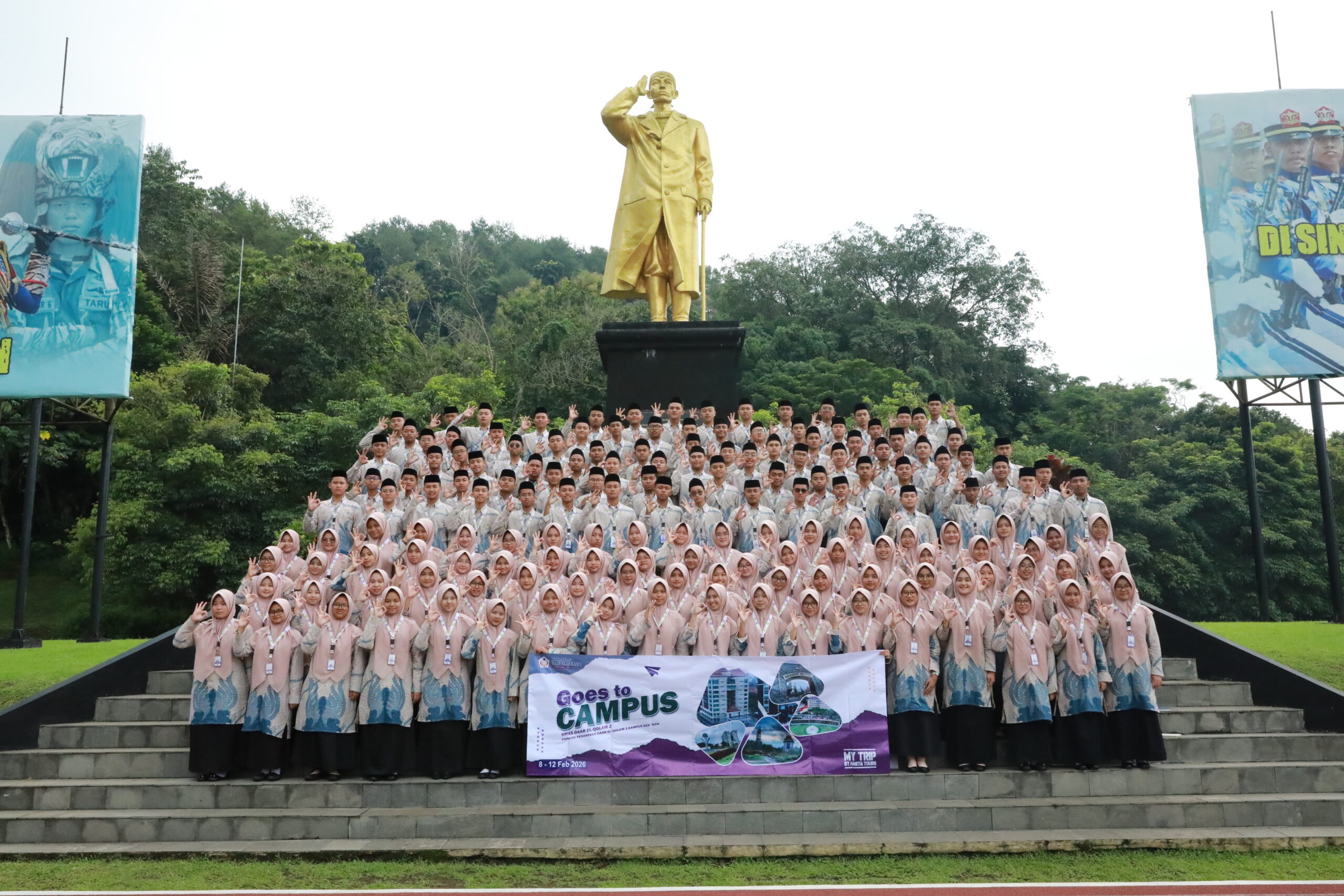 Large student group in pink-and-blue uniforms on steps before a tall golden saluting statue, holding Goes to Campus banner.