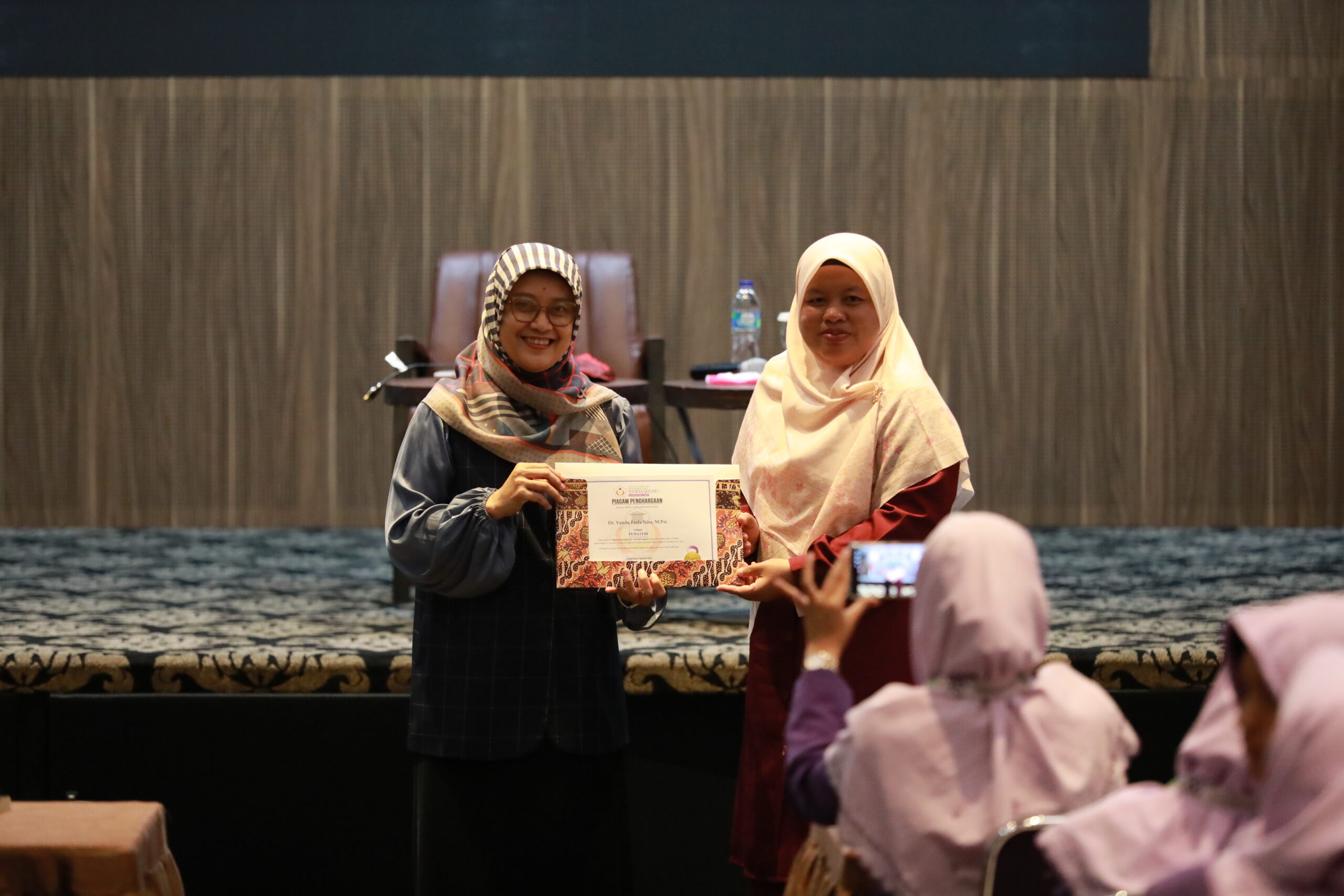 Two smiling women in hijab on stage holding a certificate together while an audience member takes a photo