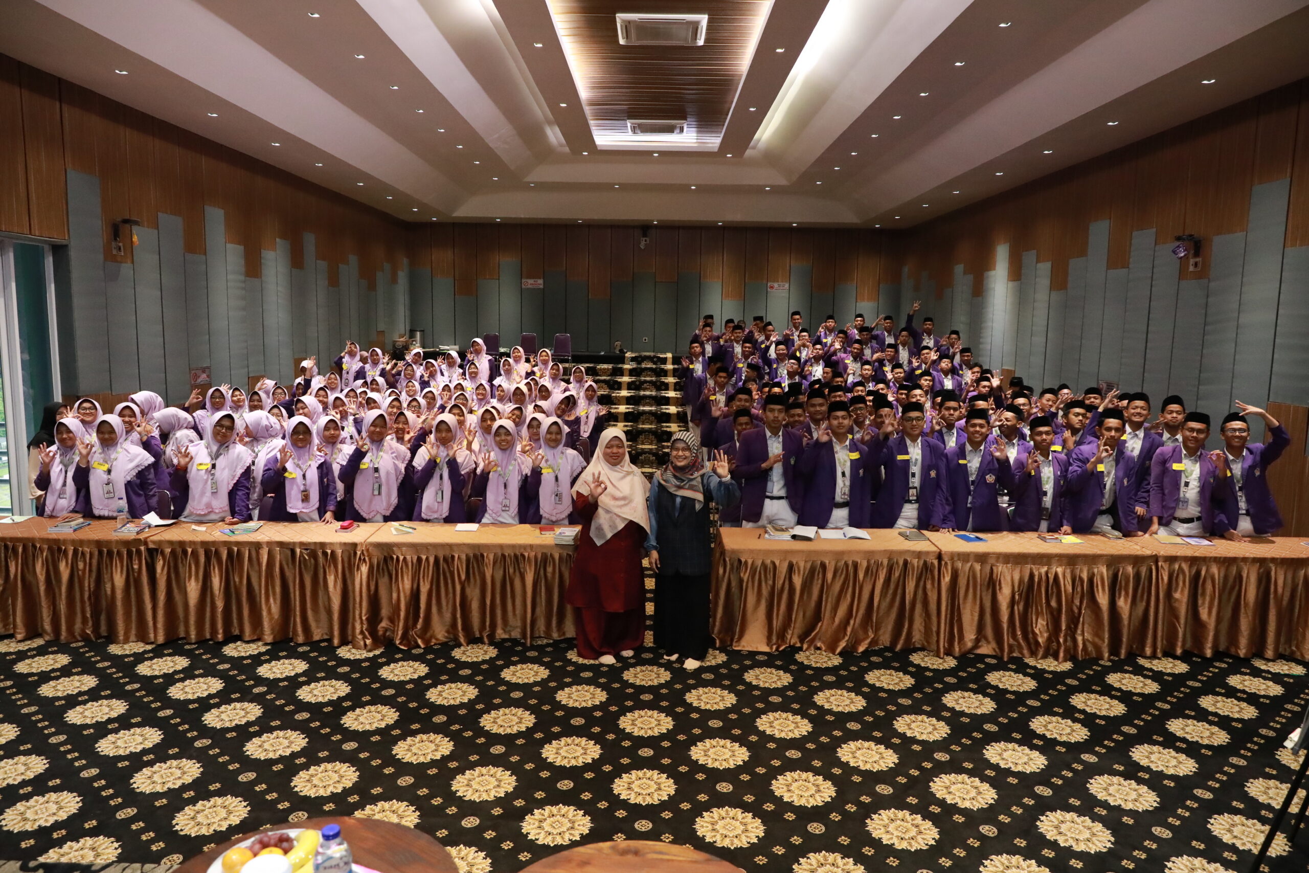 Large group of students in purple uniforms and hijabs posing in a conference room with two teachers at front.