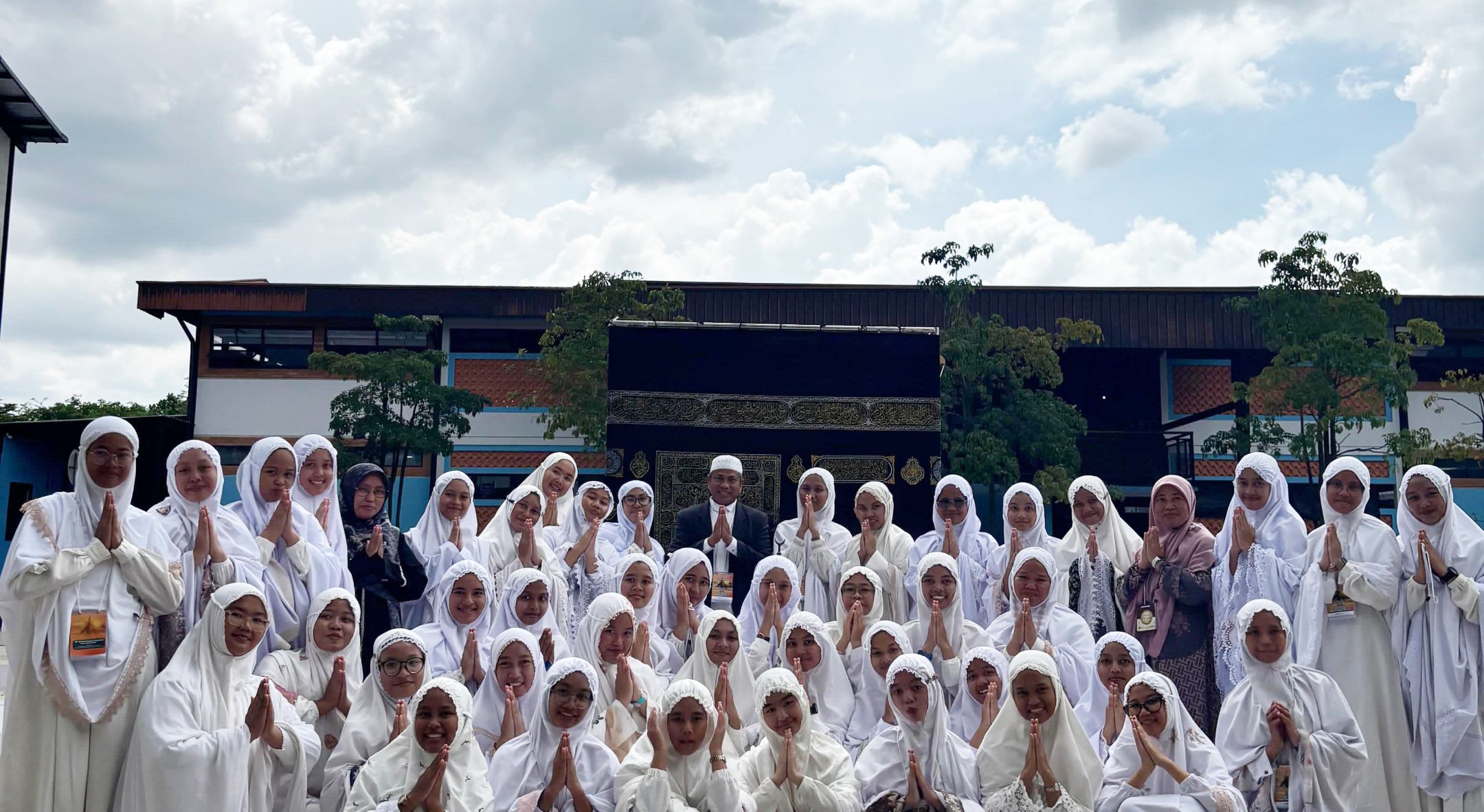 Large group of girls and women in white prayer robes posing with hands together in front of a Kaaba replica outdoors.