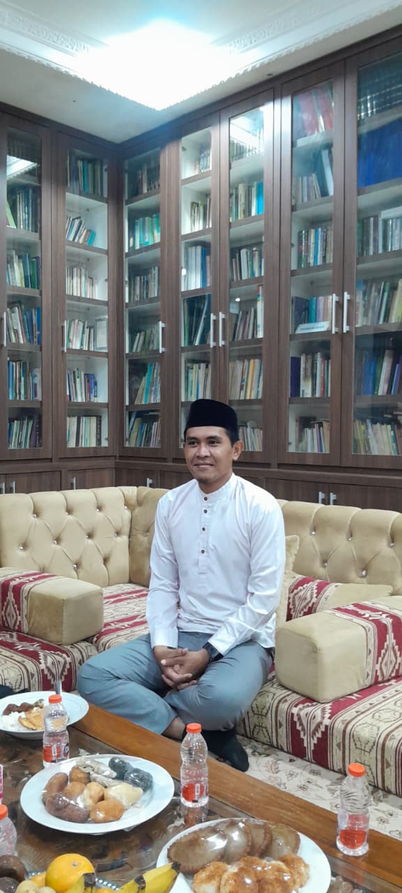 Man in white shirt and black cap sitting cross-legged on a patterned sofa in front of tall bookcases, table with snacks.