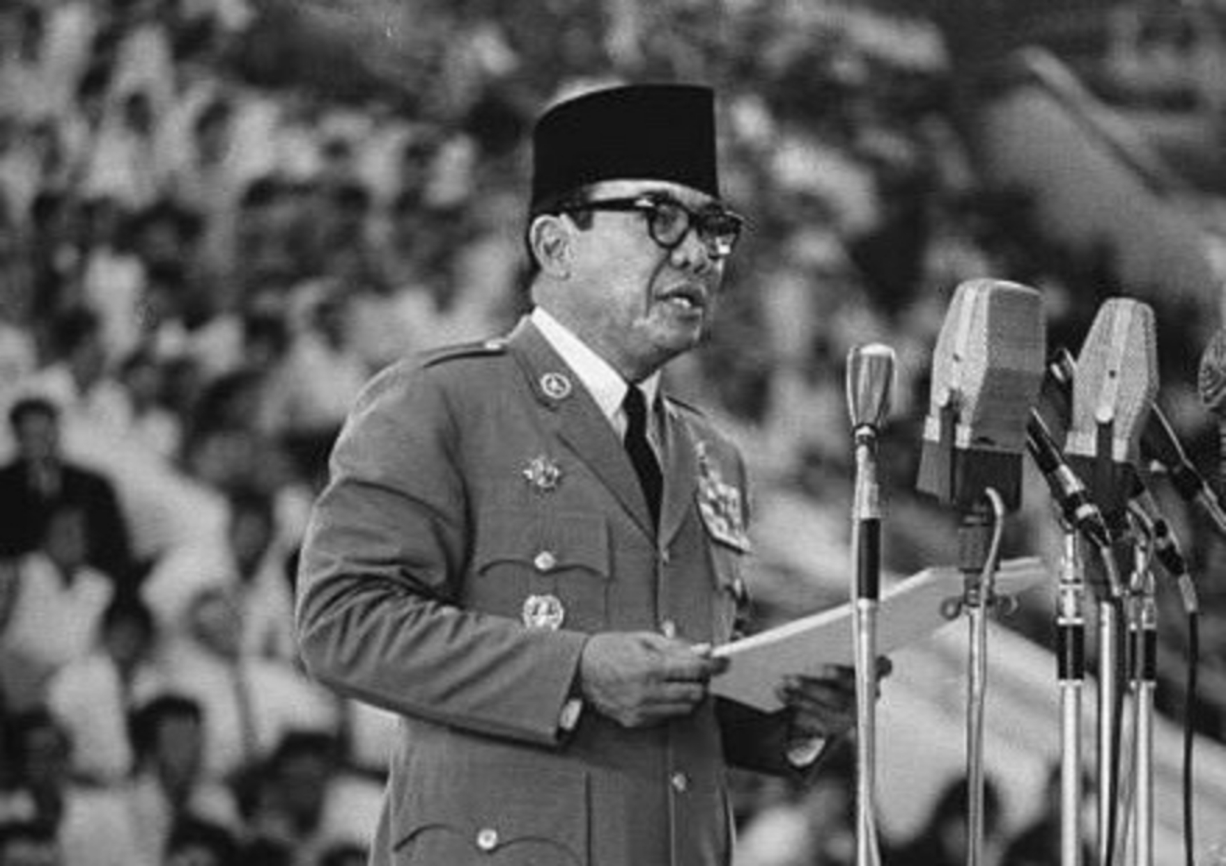 Man in decorated military uniform and black cap delivering a speech at a podium into multiple microphones, holding papers.