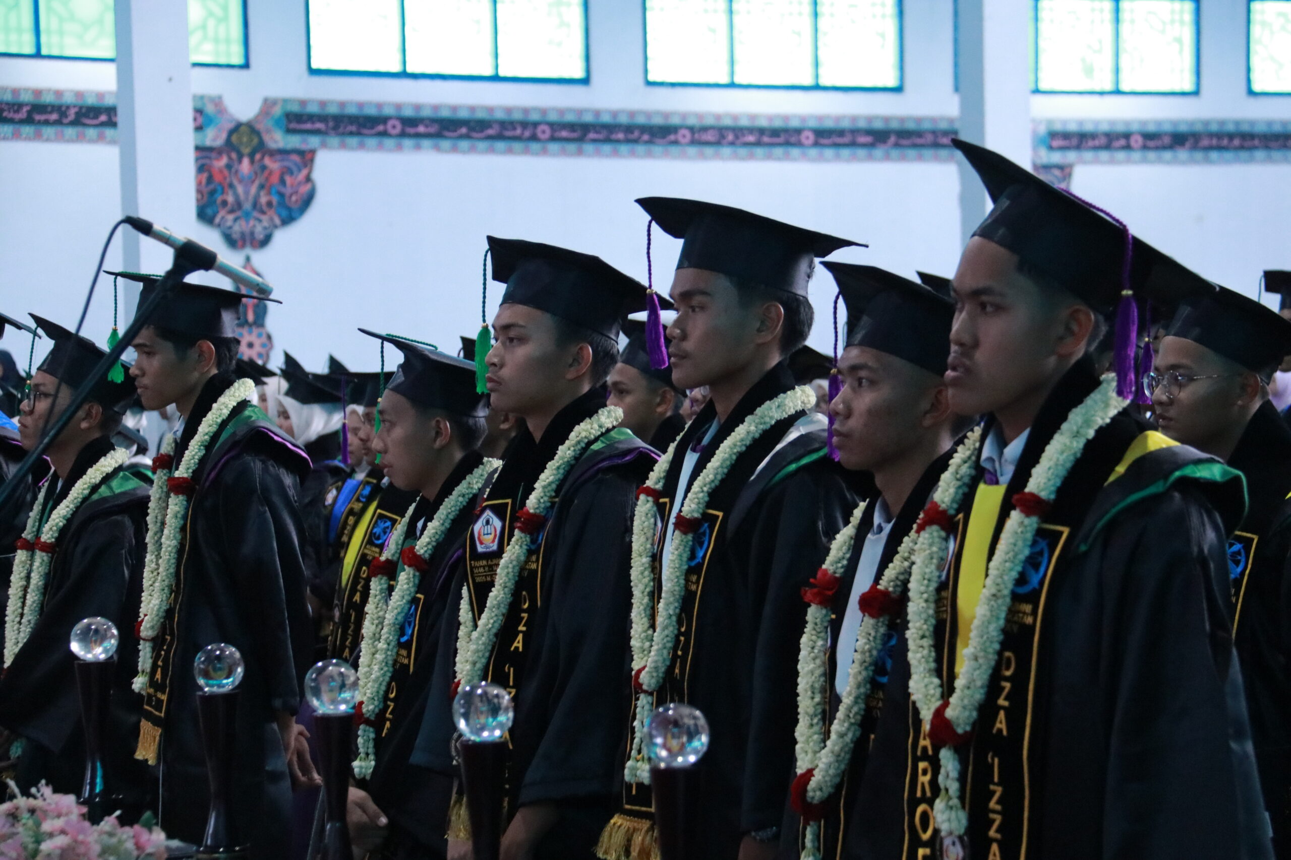 Line of graduates in mortarboard caps and gowns wearing floral garlands and stoles, standing solemnly in a decorated hall.