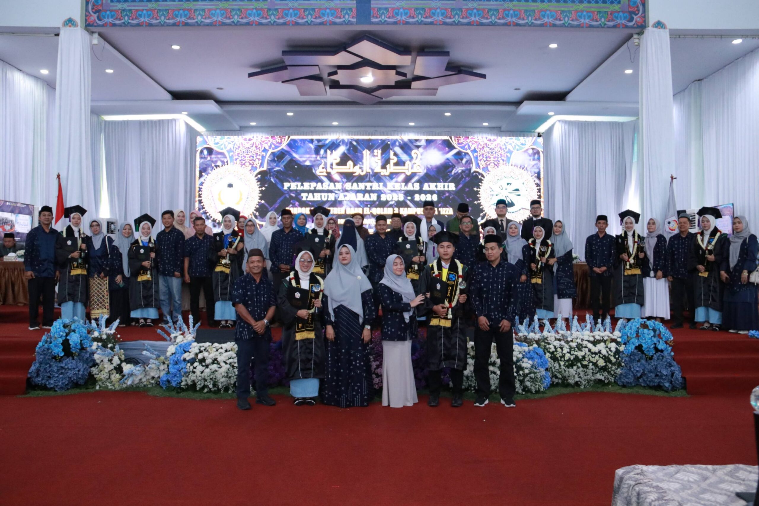 Large group of graduates and staff in dark robes and headscarves posing on a decorated stage with flowers and a banner.