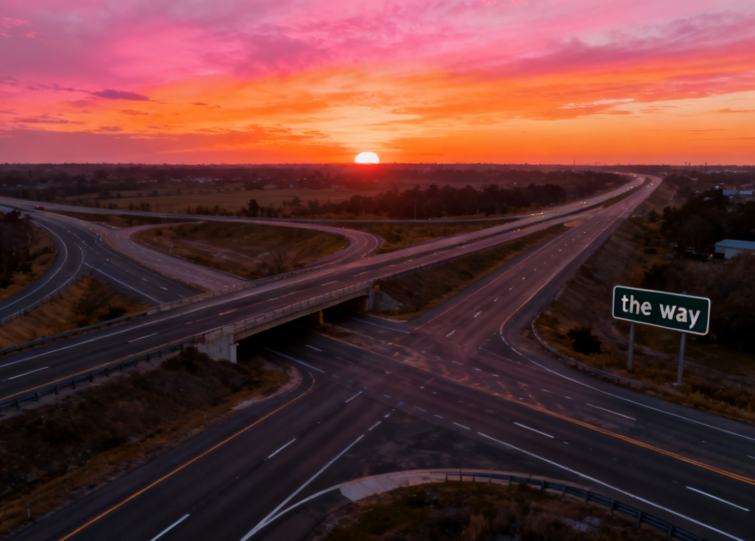Sunset over an empty multi‑lane highway interchange with looping ramps and a green roadside sign reading "the way"