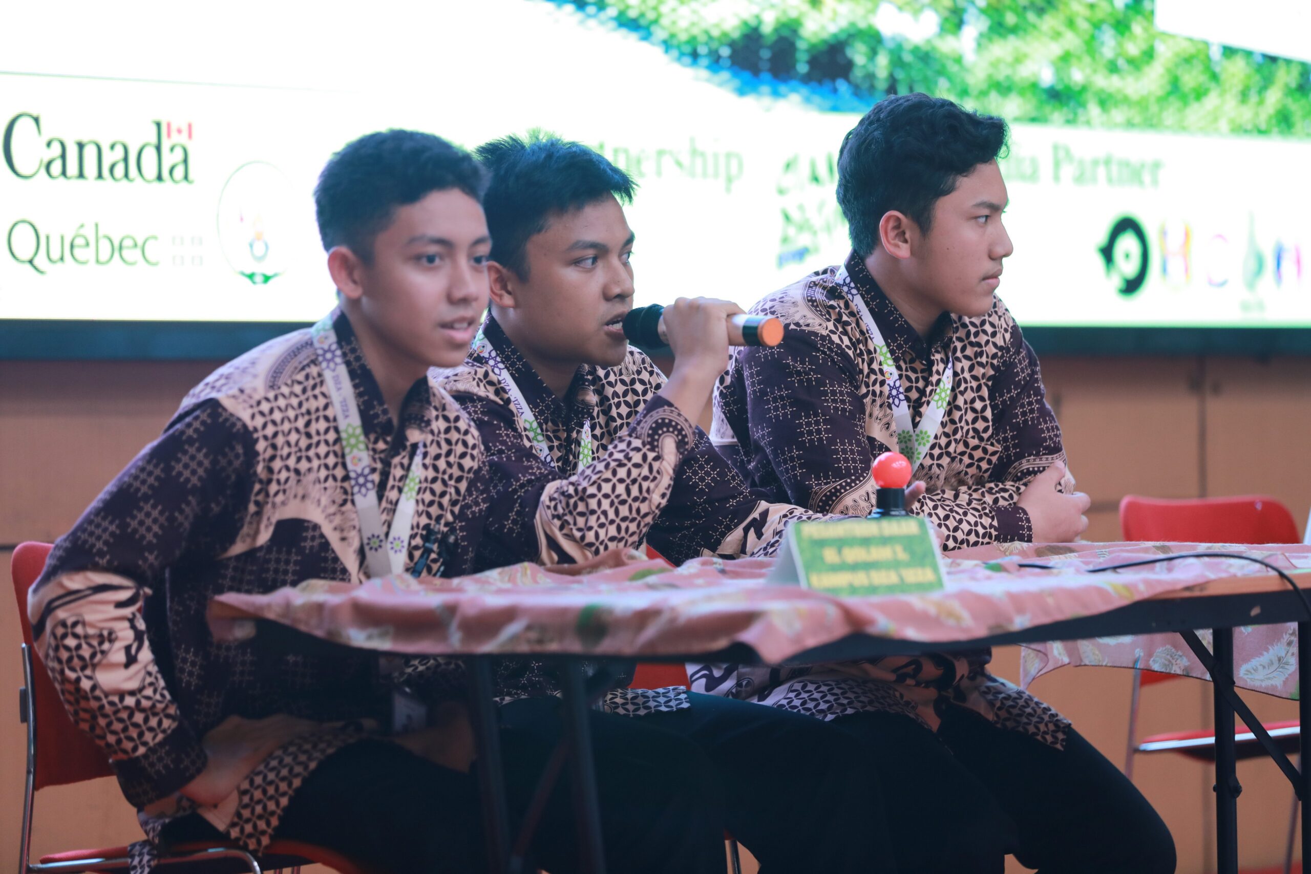 Three teenage boys in patterned shirts sit at a table; the middle boy holds a microphone while the others listen.