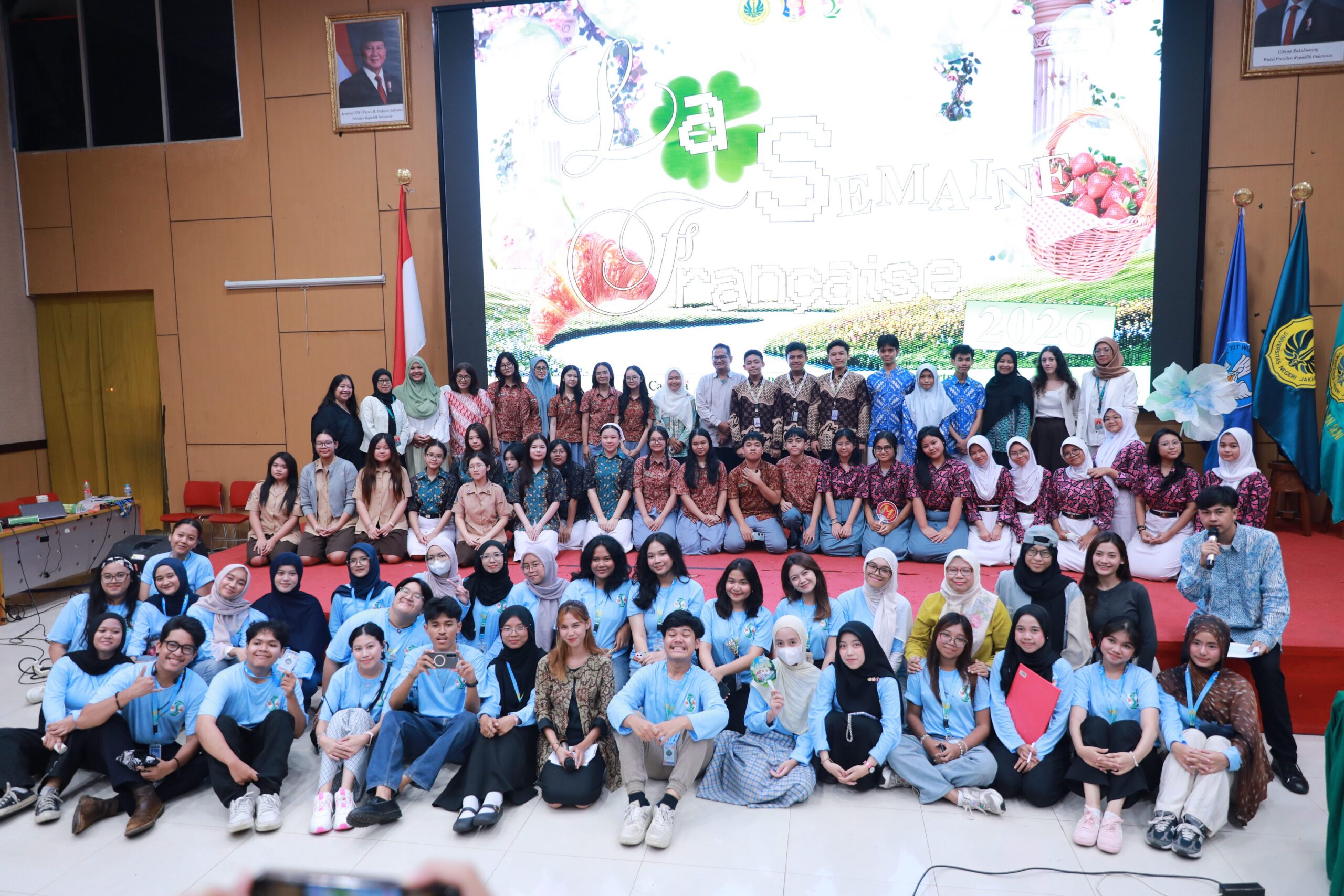 Large group of smiling students and teachers posing on stage in front of a colourful screen and flags during an event.