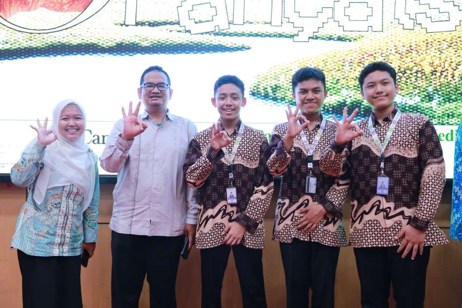 Five smiling students and two adults in patterned shirts stand in a row, each making an OK hand gesture at an event.
