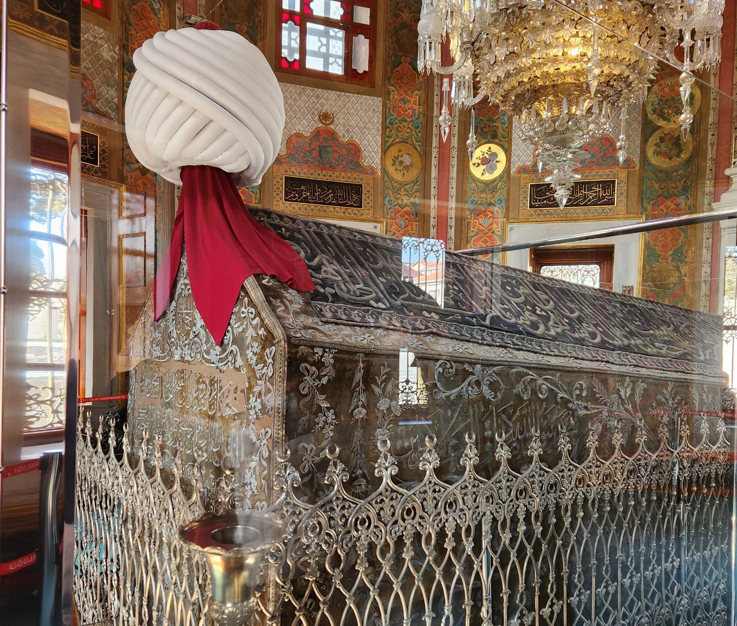 Ornate Ottoman tomb with embroidered drapery, a large white turban with red sash atop the coffin, and decorative metal grille in a richly painted chamber.