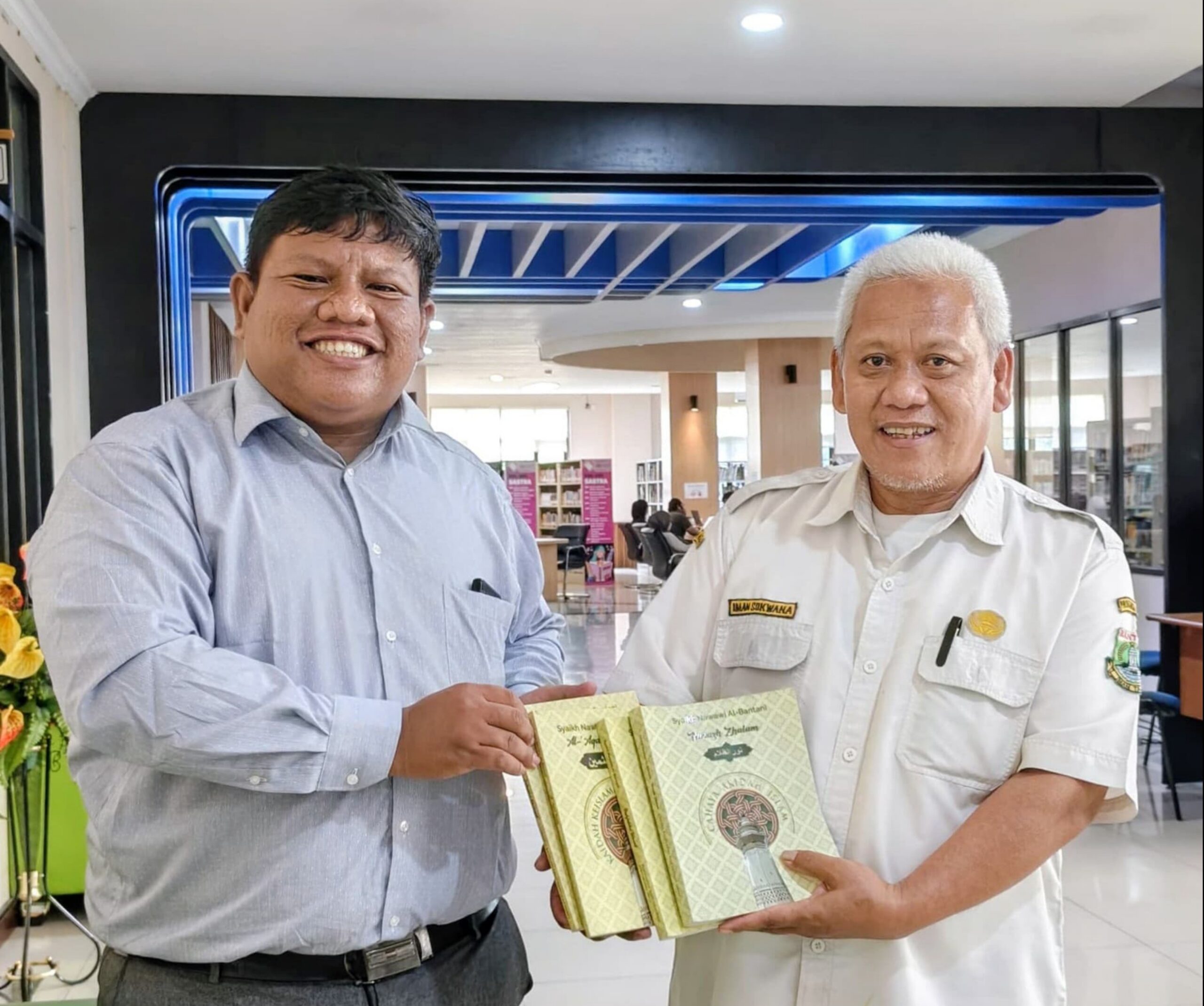 Two smiling men in shirts holding and presenting three green patterned books inside a modern library lobby.