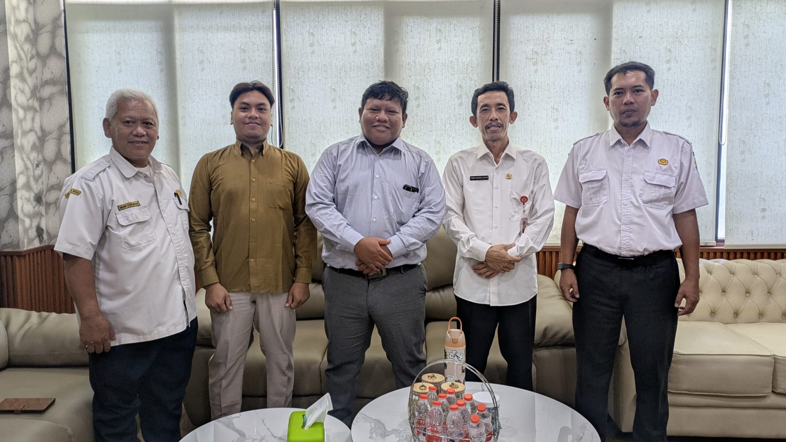 Five men standing in a row in an office lounge, dressed in shirts and trousers, smiling for a group photo.