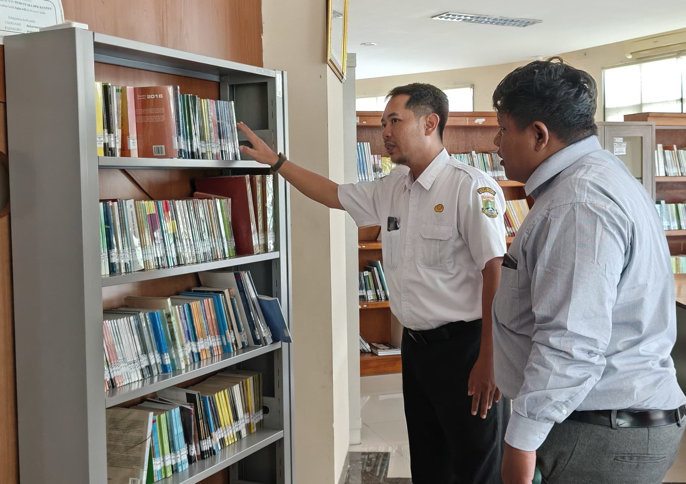 Two men in a library examining books on a tall metal shelf, one pointing toward the upper shelf while the other watches.