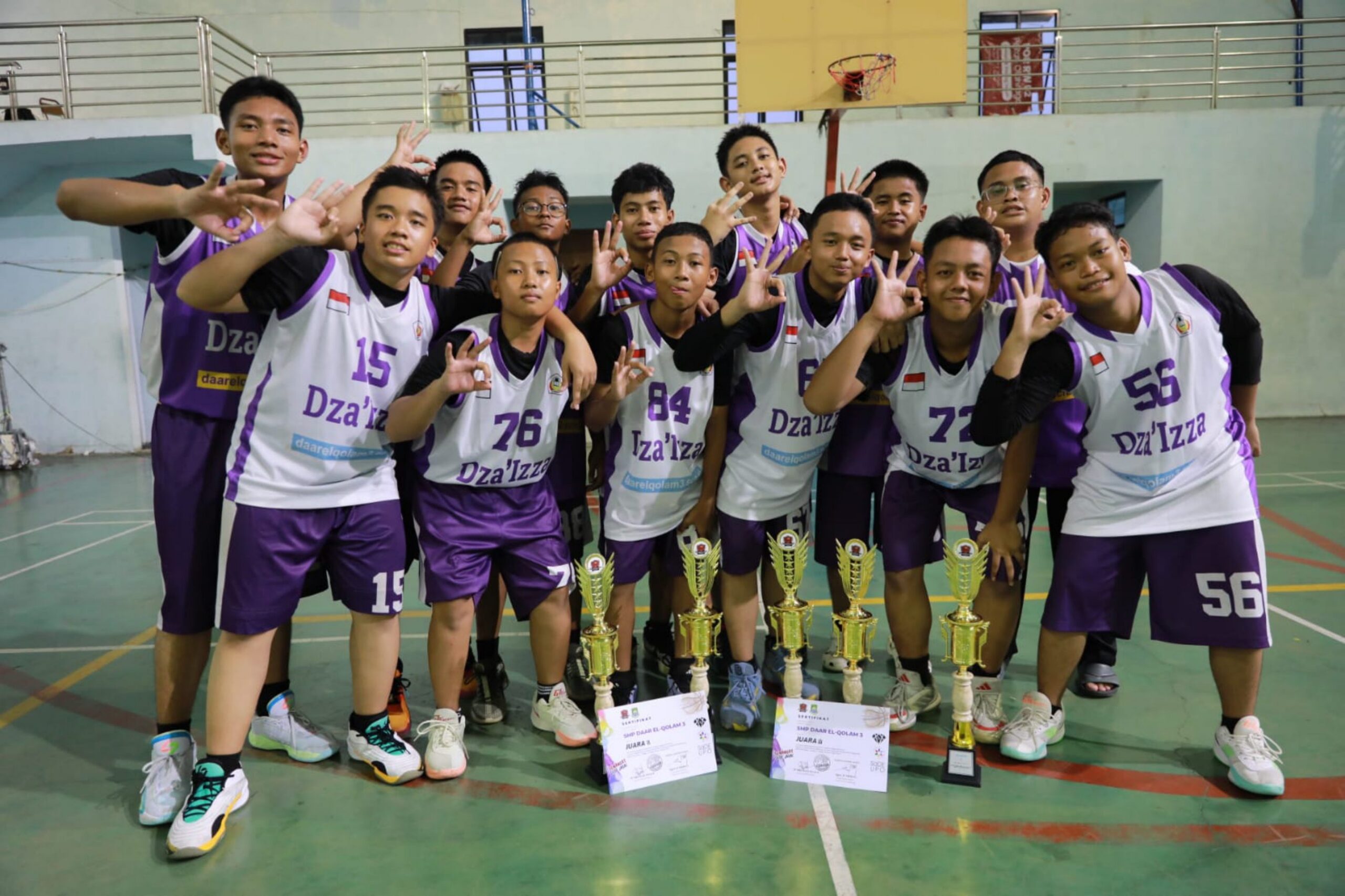 Youth basketball team in purple and white jerseys posing indoors behind trophies and certificates, many making OK hand signs.