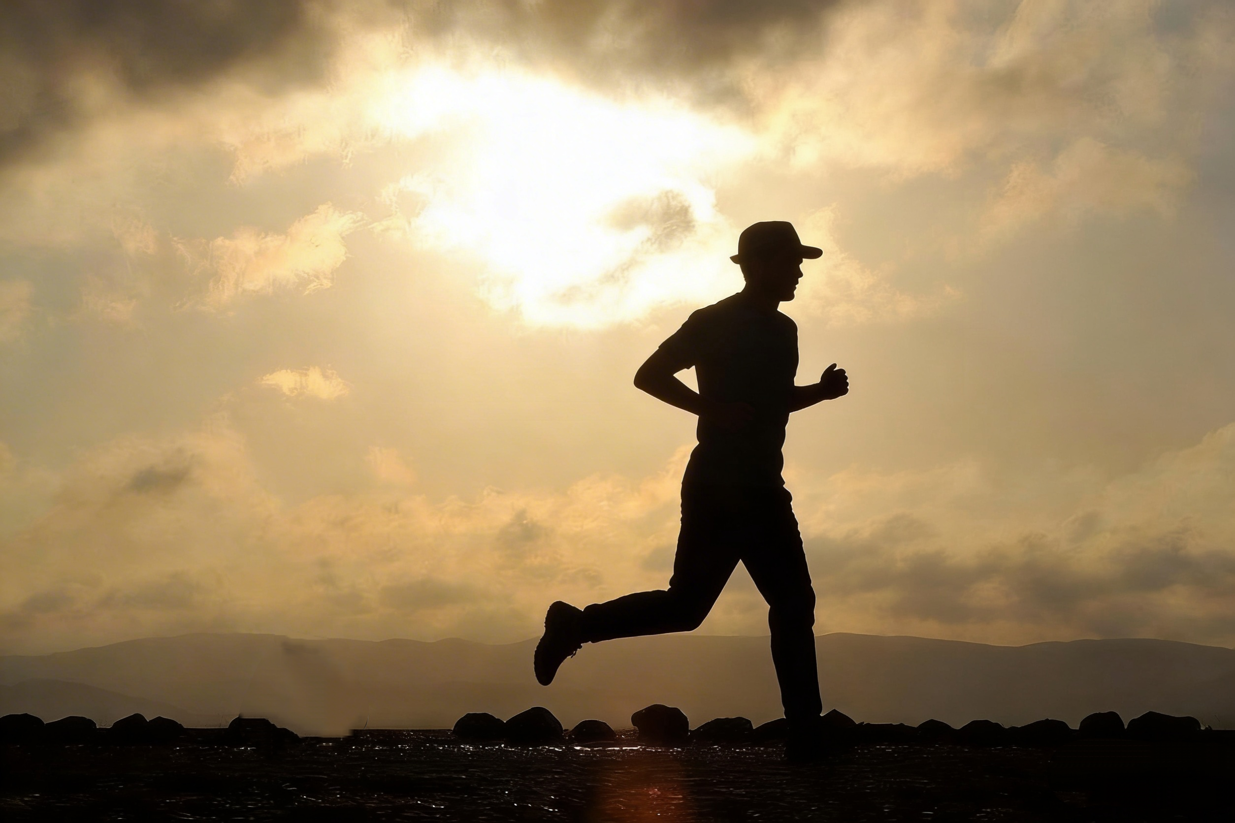 Silhouette of a person wearing a hat jogging along a rocky shoreline at sunset with a glowing cloudy sky.