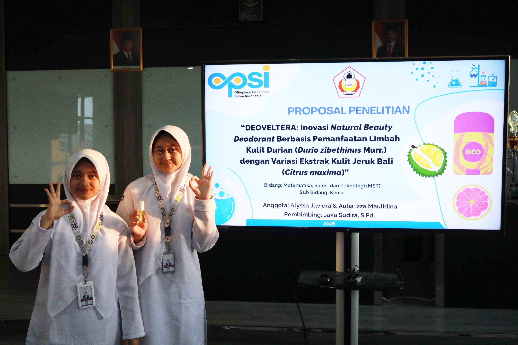 Two female students in white hijabs hold a small deodorant bottle and make OK signs in front of a research proposal screen.